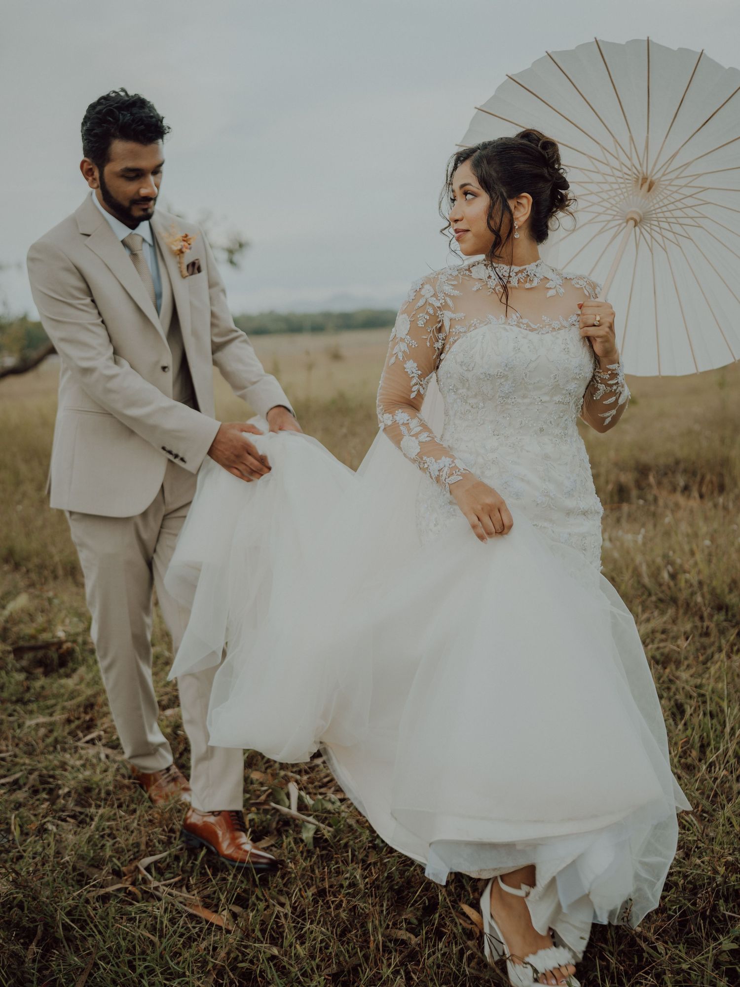 Bride in a lace long-sleeve wedding gown holding a white parasol walks beside a groom in a beige suit across a grassy field.