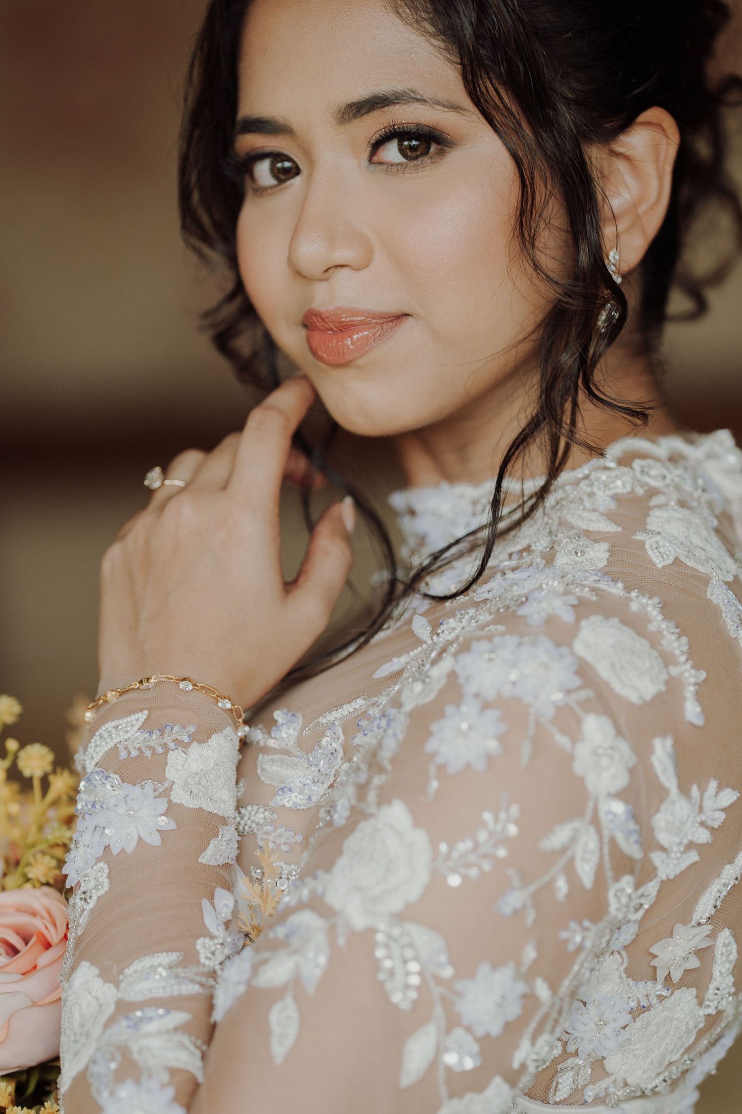 Close-up portrait of a smiling bride in a sheer floral lace gown, hand raised to her face, with an orange rose bouquet beside her.