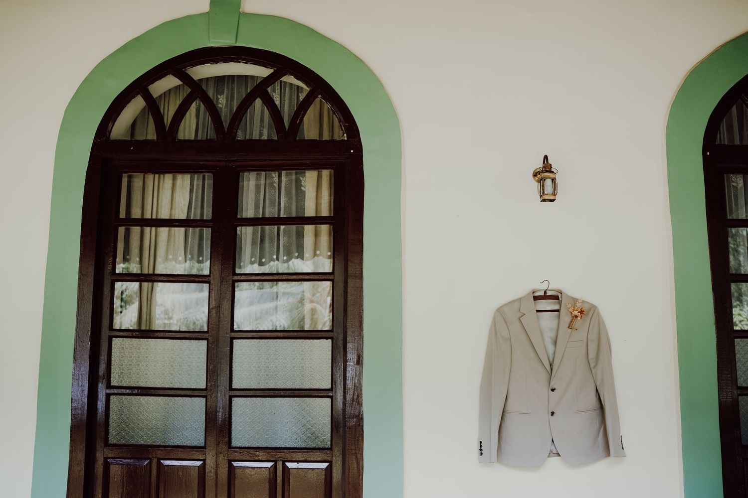 Beige groom's suit jacket with a boutonniere hangs on a white wall beside an arched green-trimmed wooden window with glass panes.