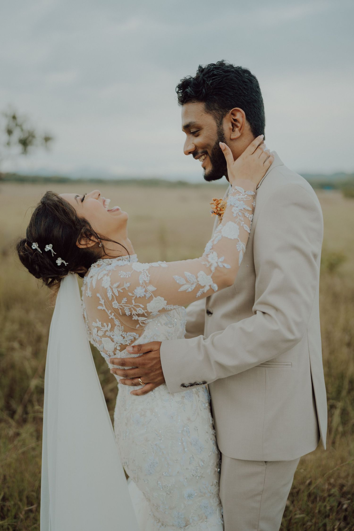A joyful bride in a lace gown and veil laughs with her head tilted back as her groom in a beige suit embraces her in a field.