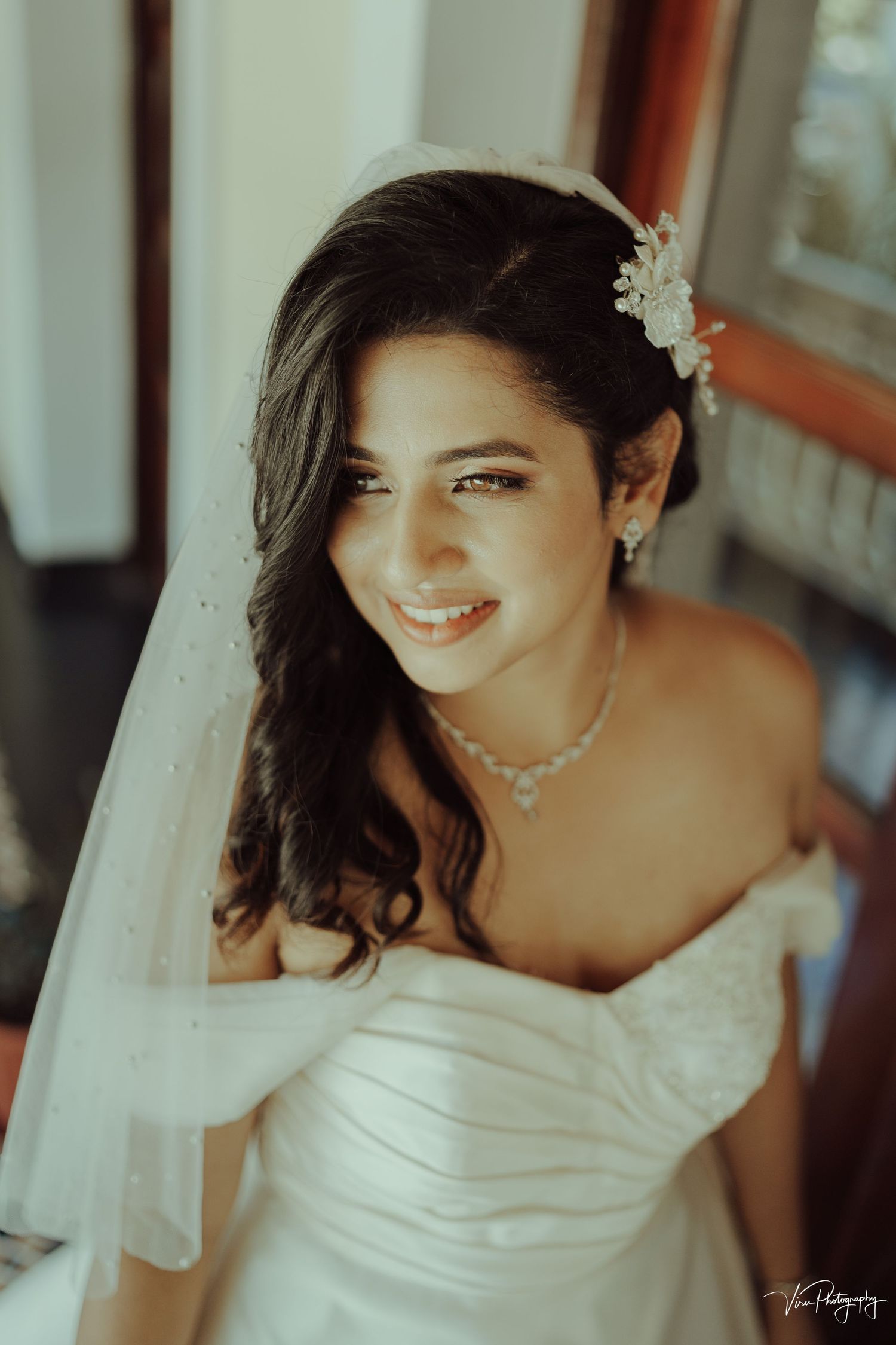 Bride wearing strapless ruched wedding gown and delicate jewelry smiles warmly in romantic natural lighting.