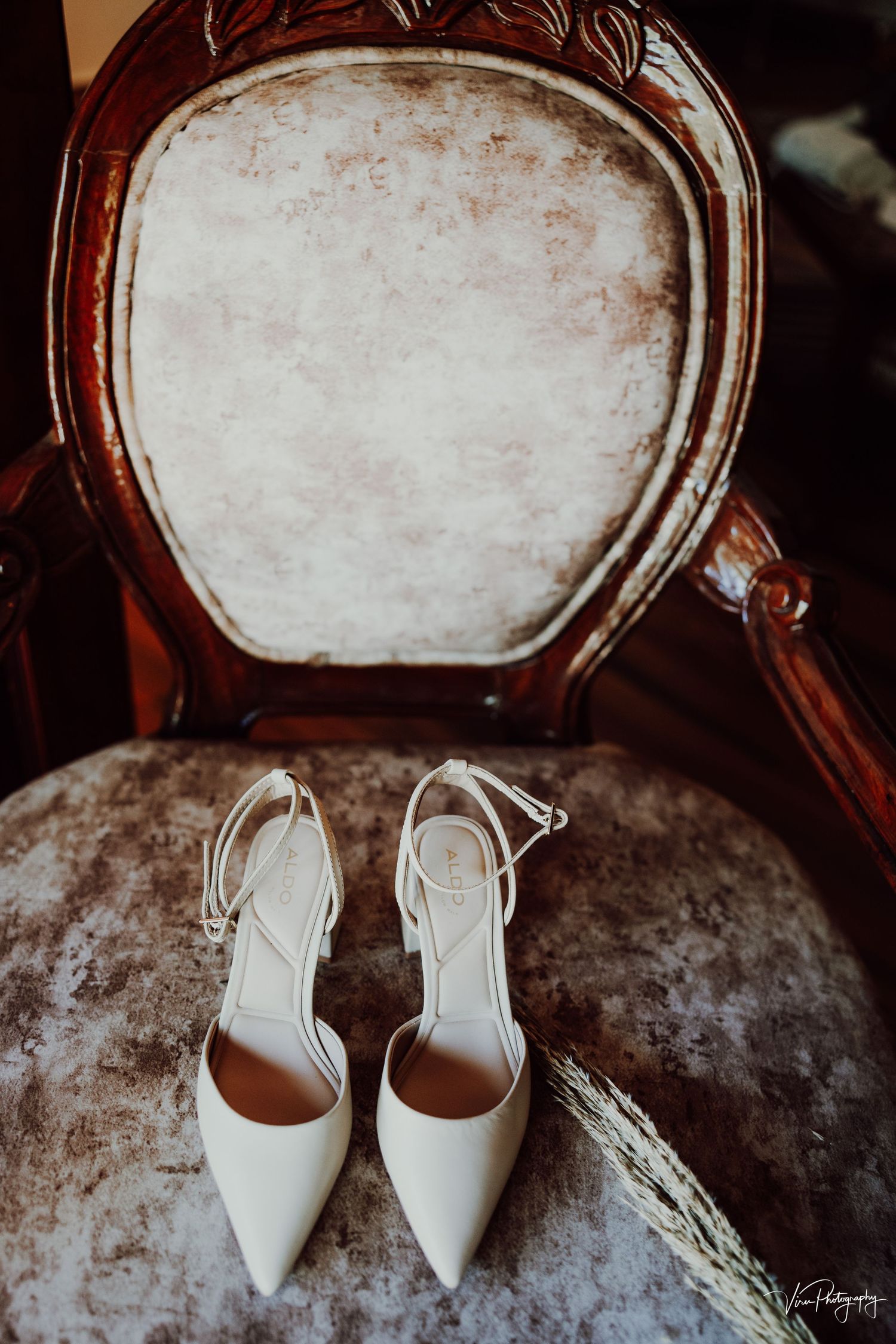 Elegant white bridal heels displayed on a vintage velvet chair with ornate wooden frame.