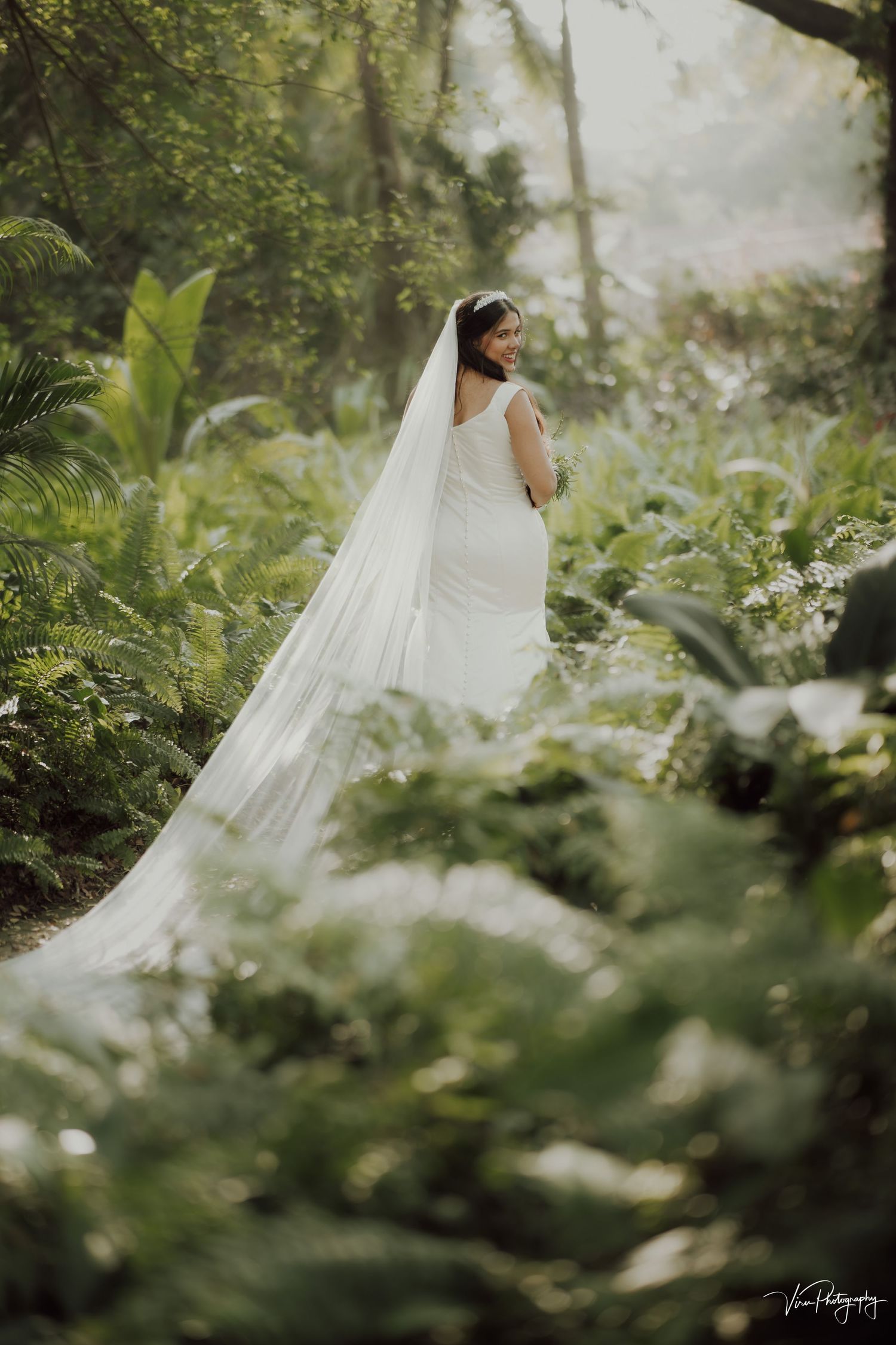 A bride in a white dress and flowing veil stands in a misty tropical garden surrounded by lush greenery and ferns.