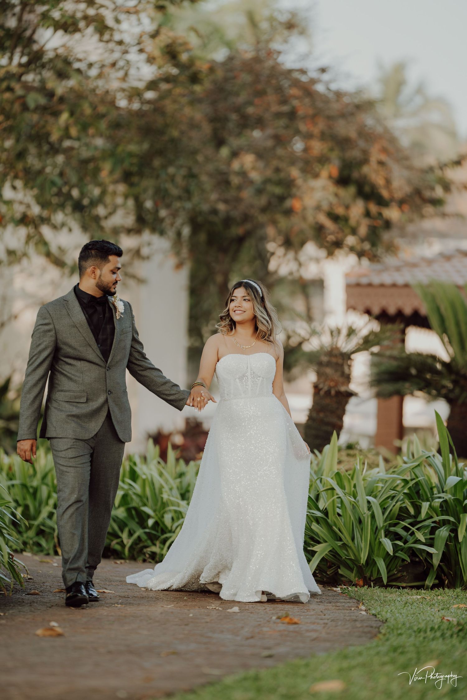 A couple in wedding attire walk along a garden path surrounded by lush greenery and palm trees at sunset.