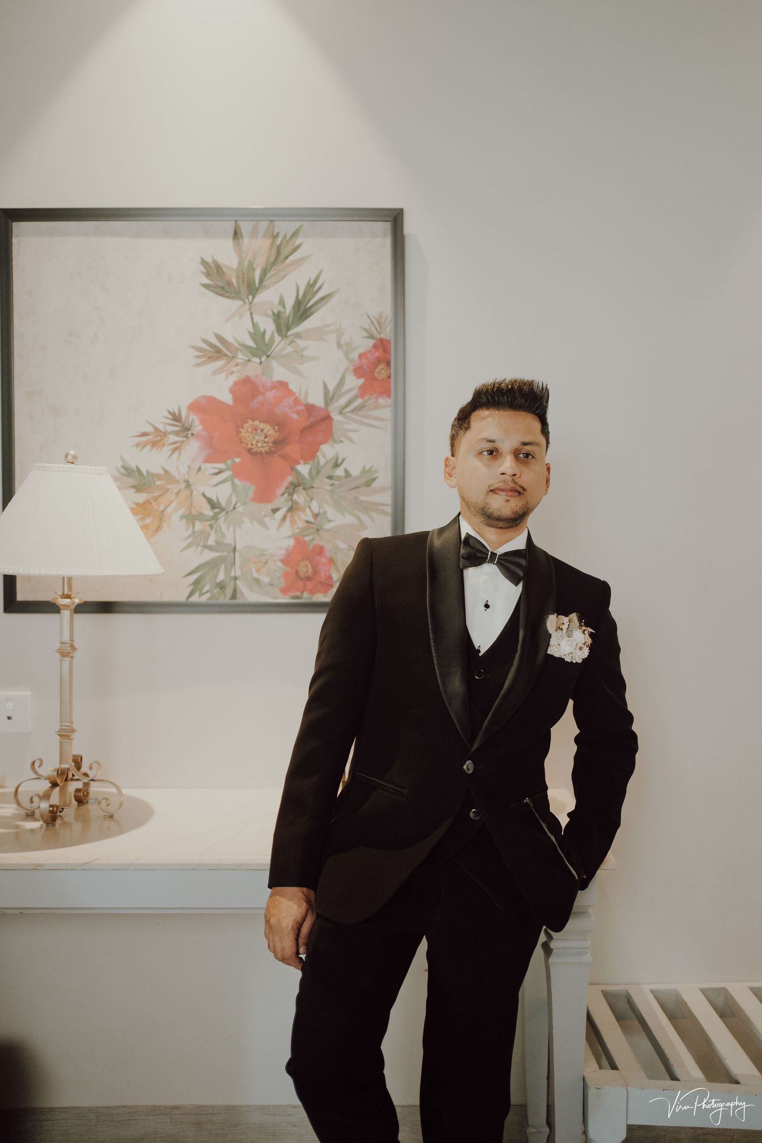 A formal black tuxedo with bow tie and boutonniere stands out against a light wall with floral artwork and candle decor.