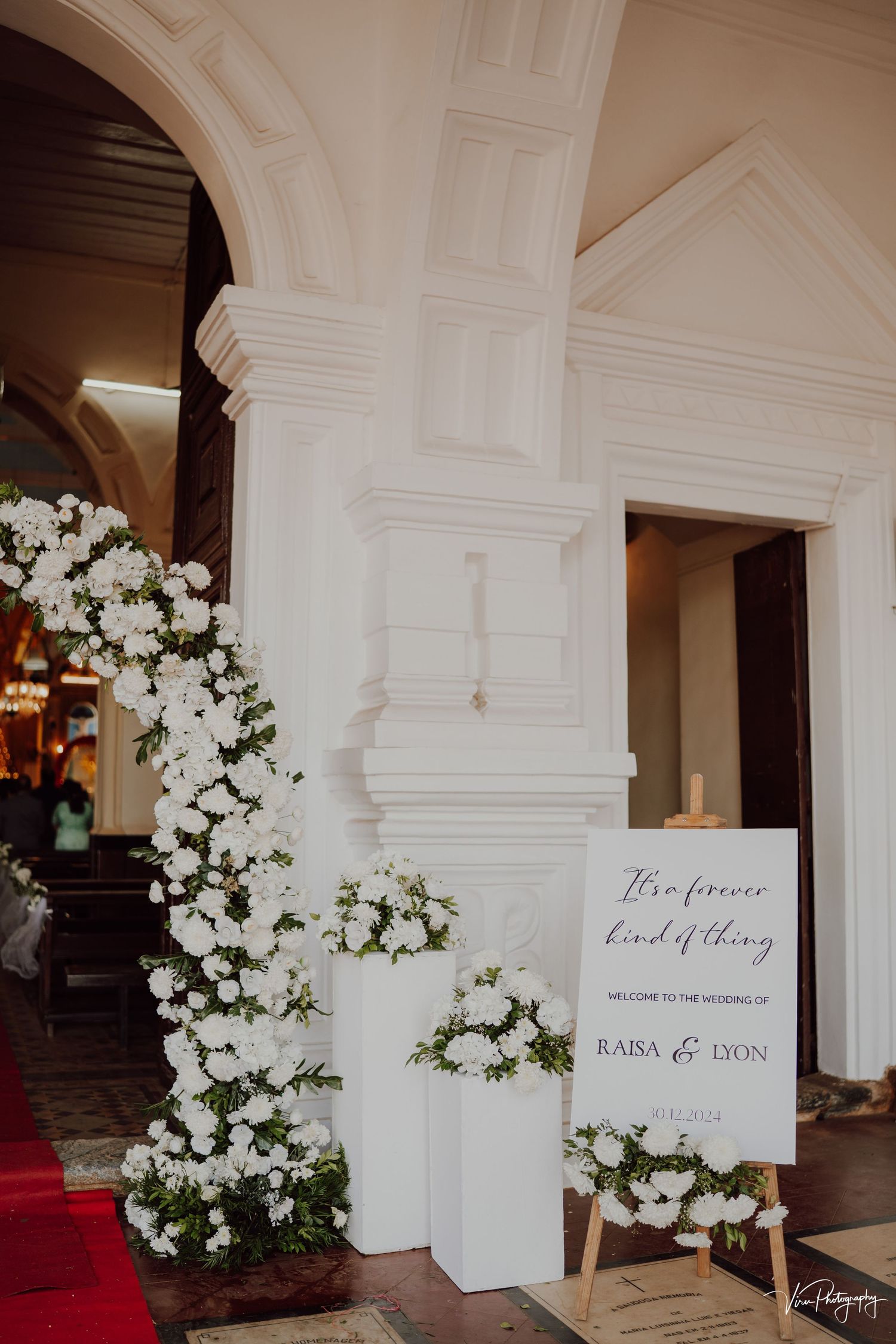 Elegant white floral arrangements and wedding signage decorate an ornate archway entrance with a red carpet.