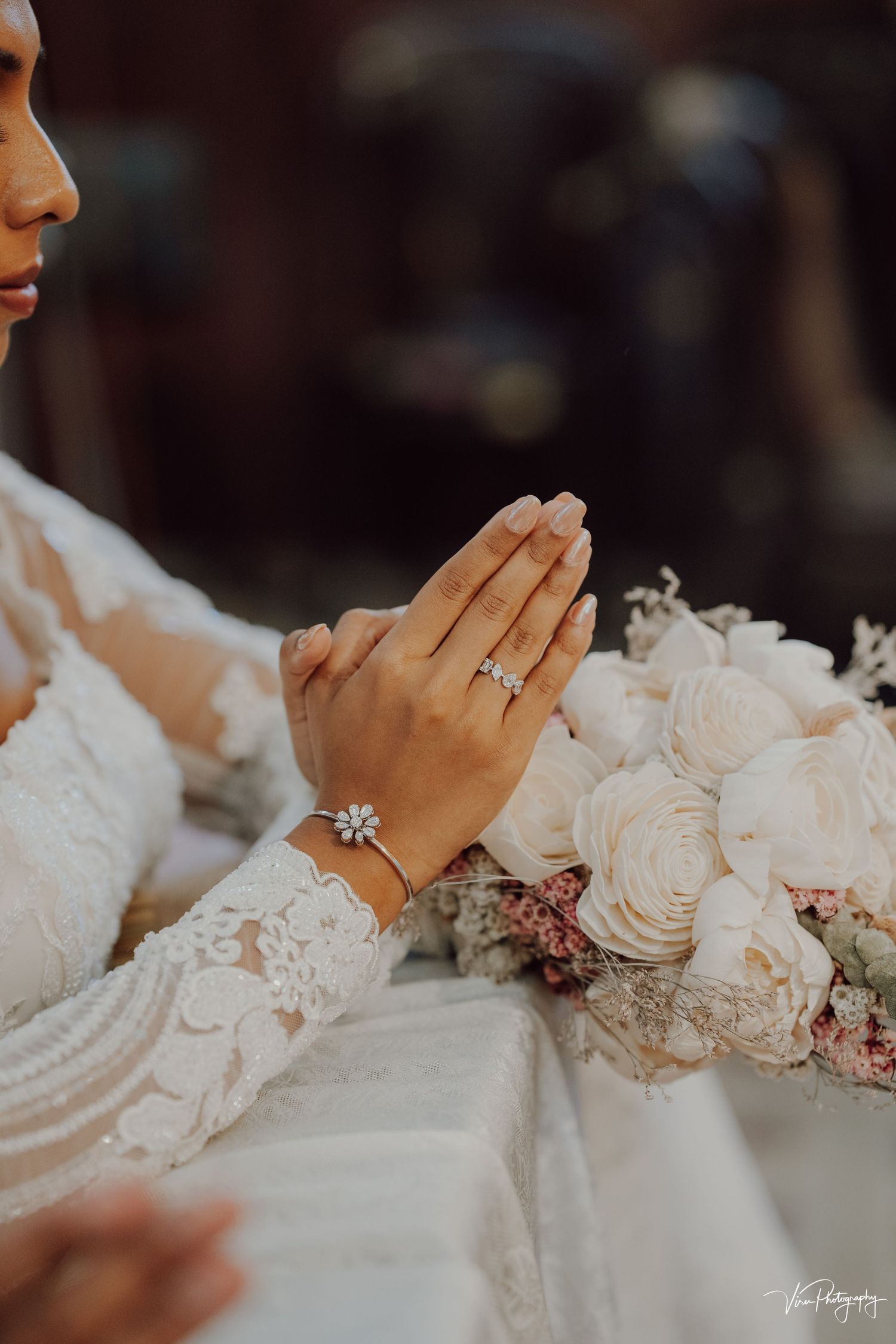 Hands clasped in prayer wearing lace sleeves and engagement ring next to white rose bouquet in soft lighting.