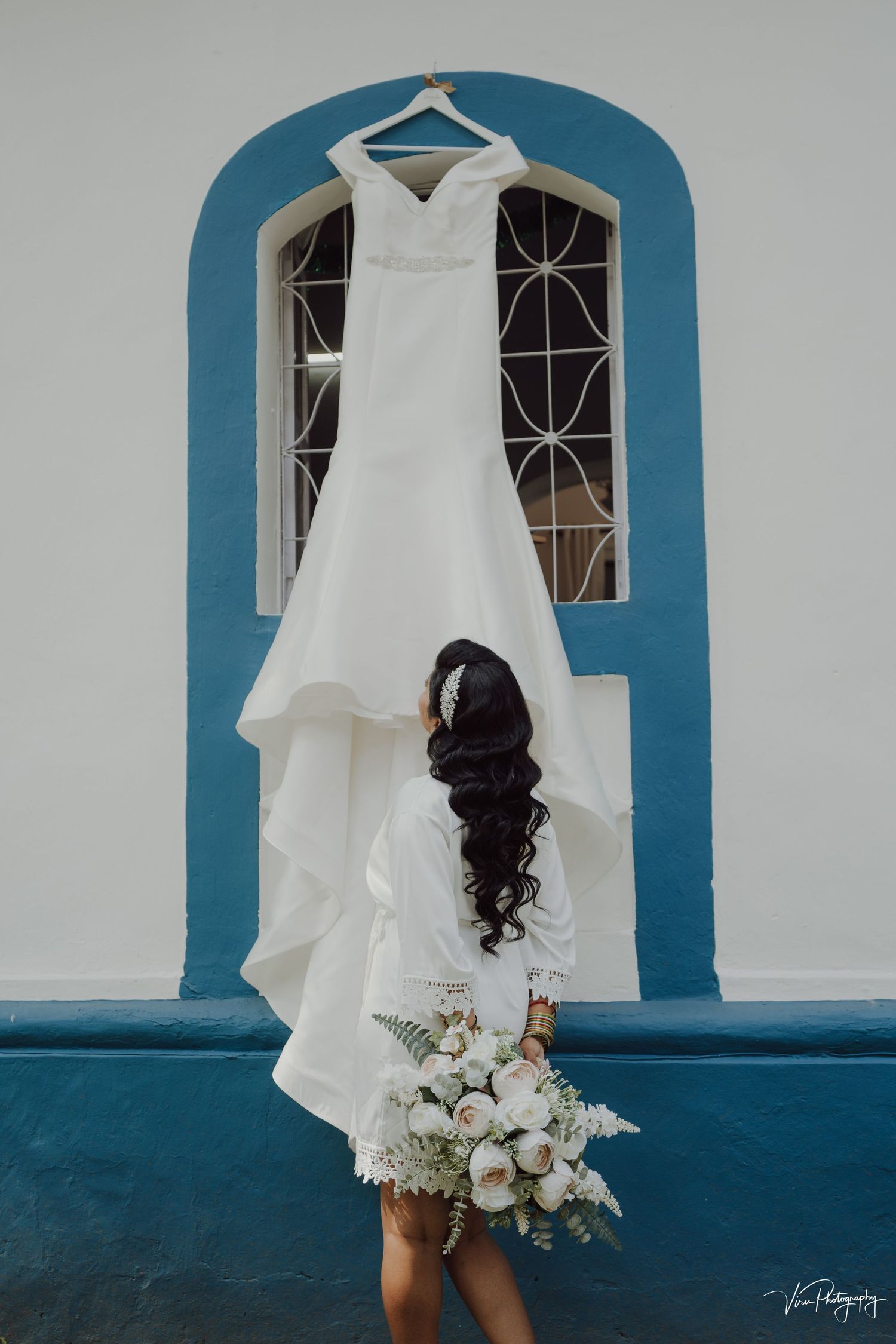 Elegant white wedding dress hangs in blue archway as person below holds beautiful white floral bouquet.