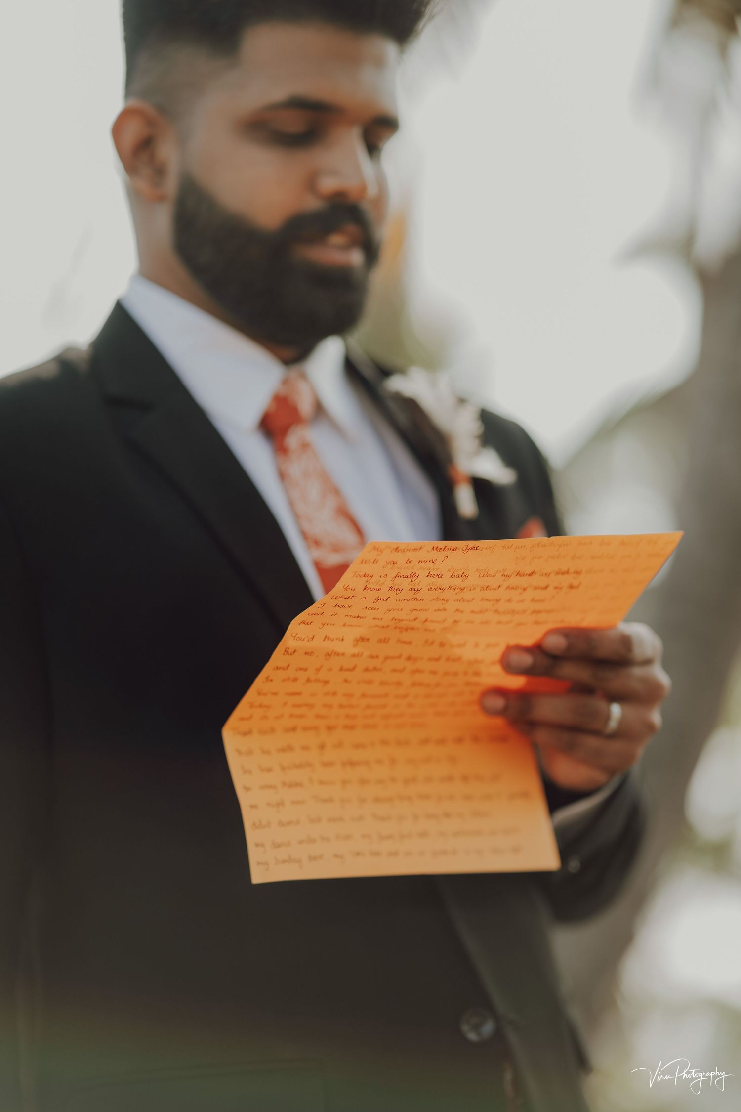A groom in a dark suit and orange tie reads from an orange paper during what appears to be a wedding ceremony.