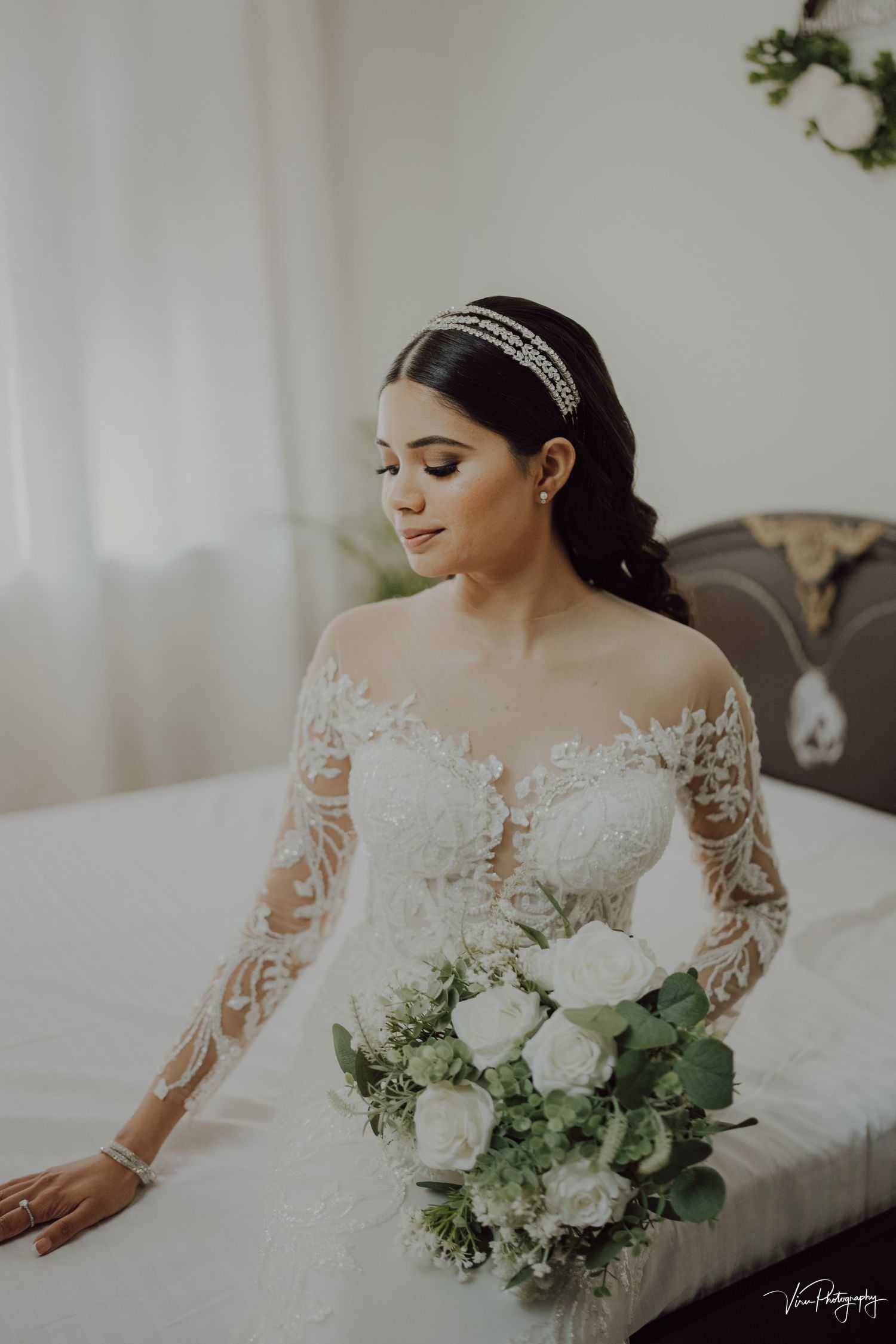 Bride in elegant lace sleeve wedding dress with sparkly headband holds white rose bouquet while sitting in bridal suite.