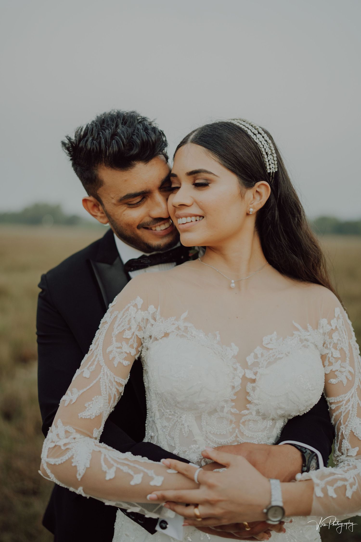 Newlywed couple embracing in outdoor field wearing formal wedding attire at golden hour.