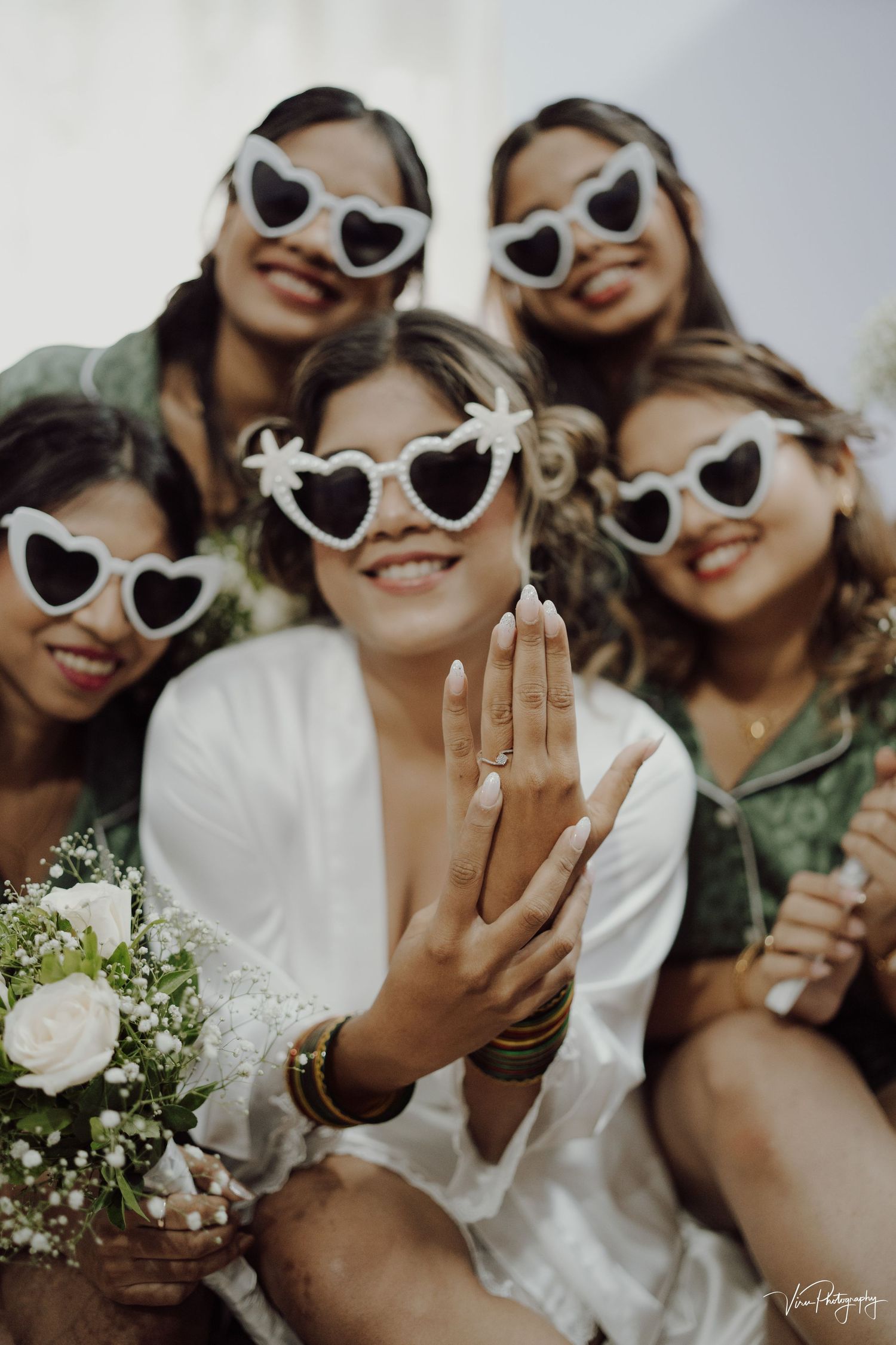 Group wearing matching heart-shaped sunglasses poses together at wedding celebration as bride shows engagement ring.