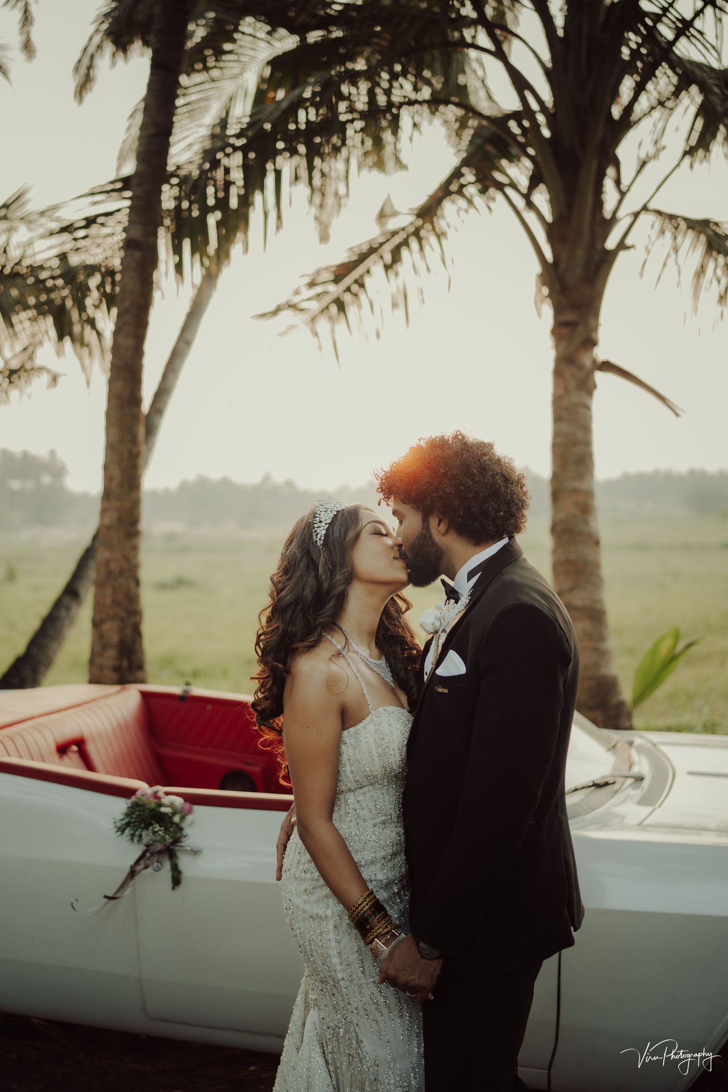 A romantic wedding couple embraces near palm trees with a vintage white car with red seats visible in the background.
