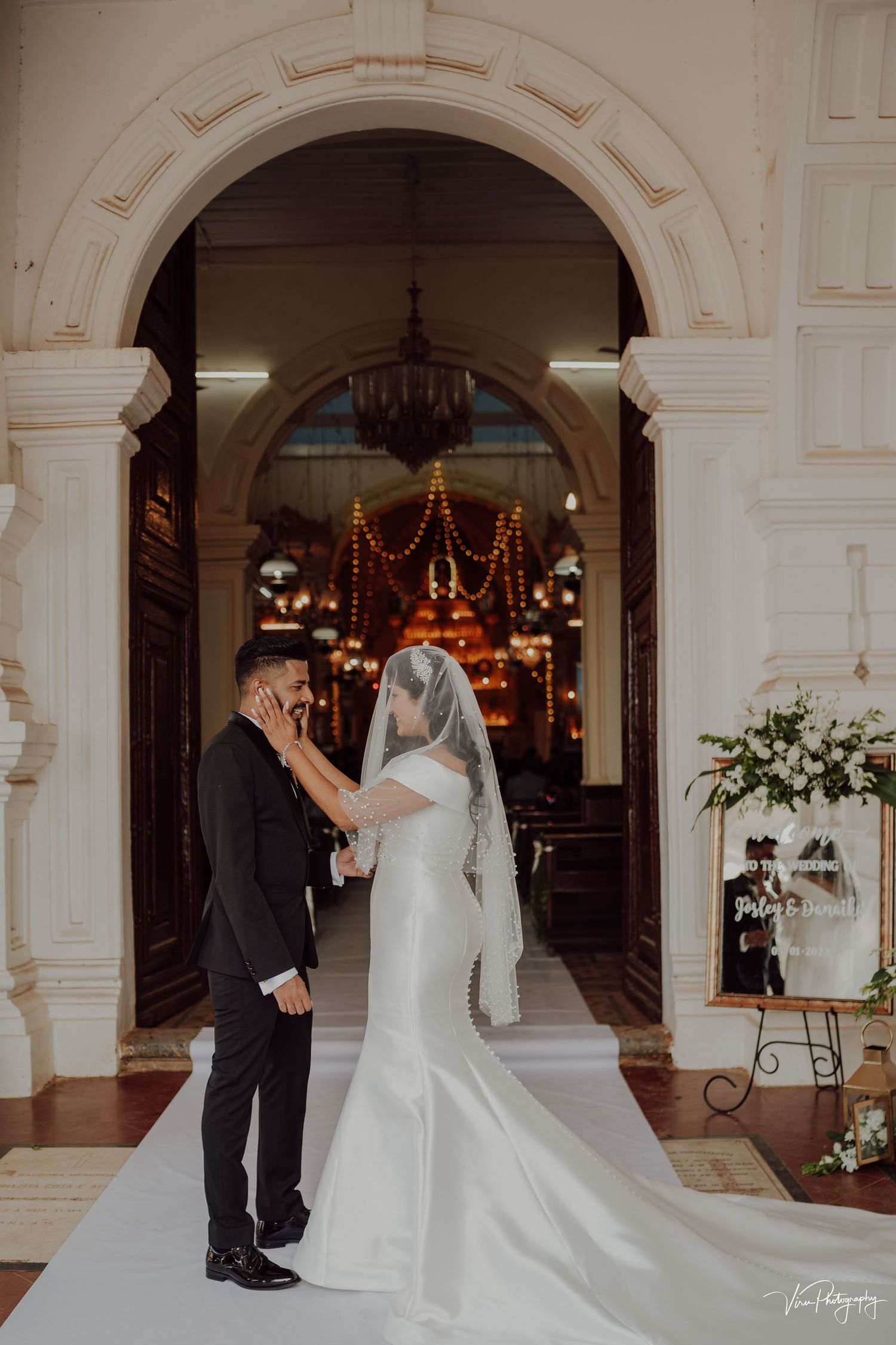 Elegant wedding ceremony takes place under a grand archway with decorative lighting and white floral arrangements.