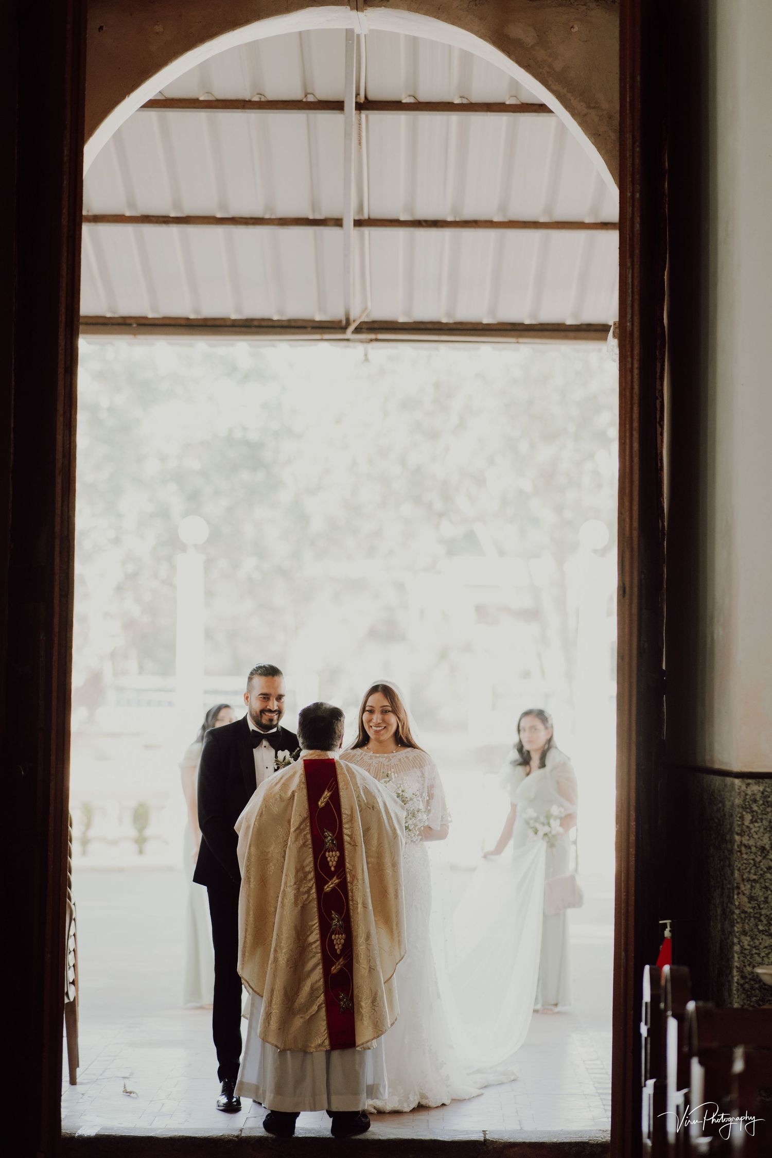 A wedding ceremony scene framed through an arched doorway with a bride in white and officiant in ceremonial robes.