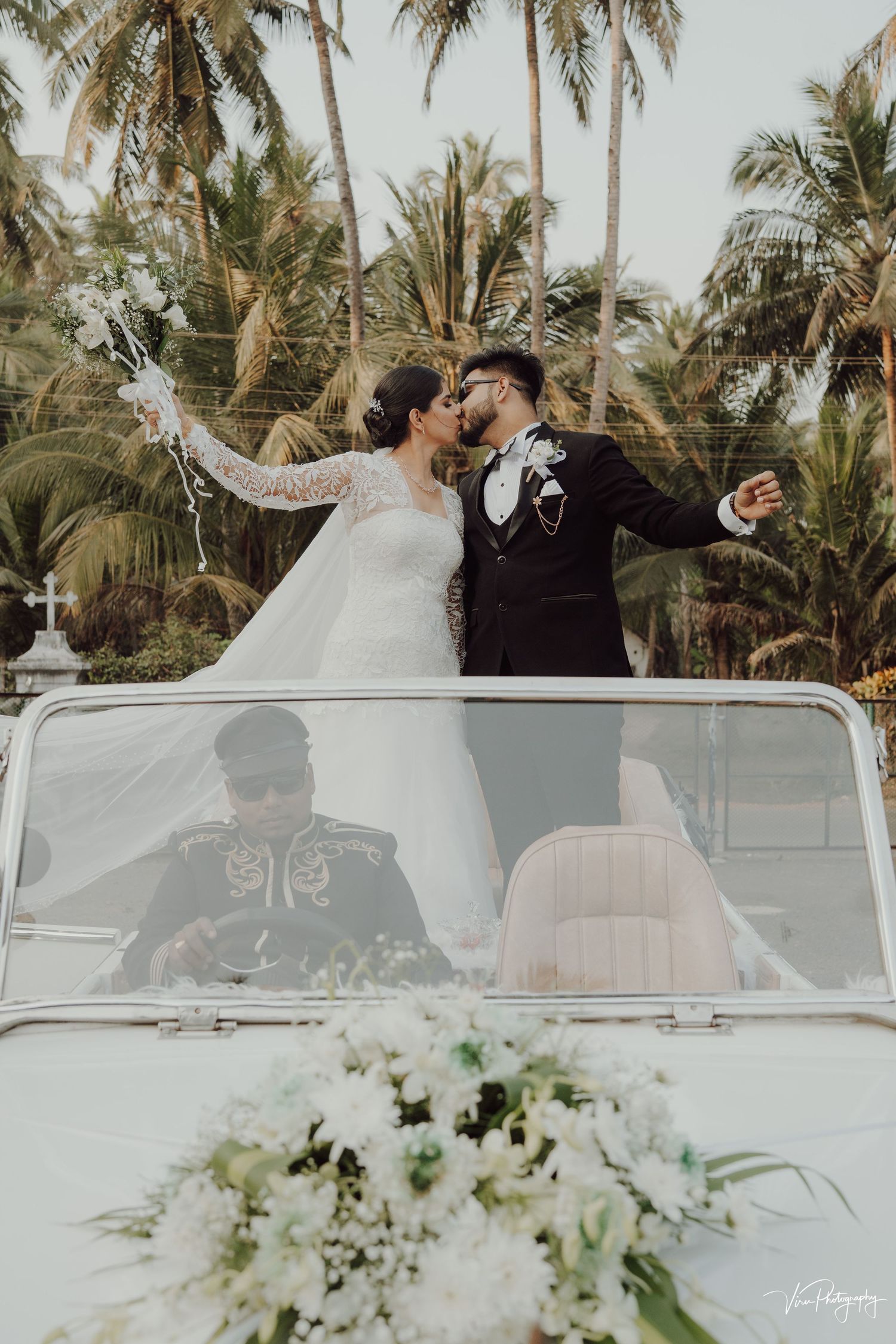 A wedding couple celebrates joyfully in a convertible decorated with white flowers among tropical palm trees.