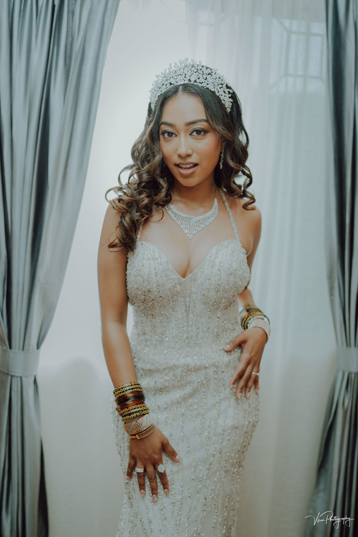Elegant bride wearing a detailed white lace wedding gown with sparkly tiara, necklace and gold bangles against curtained backdrop.