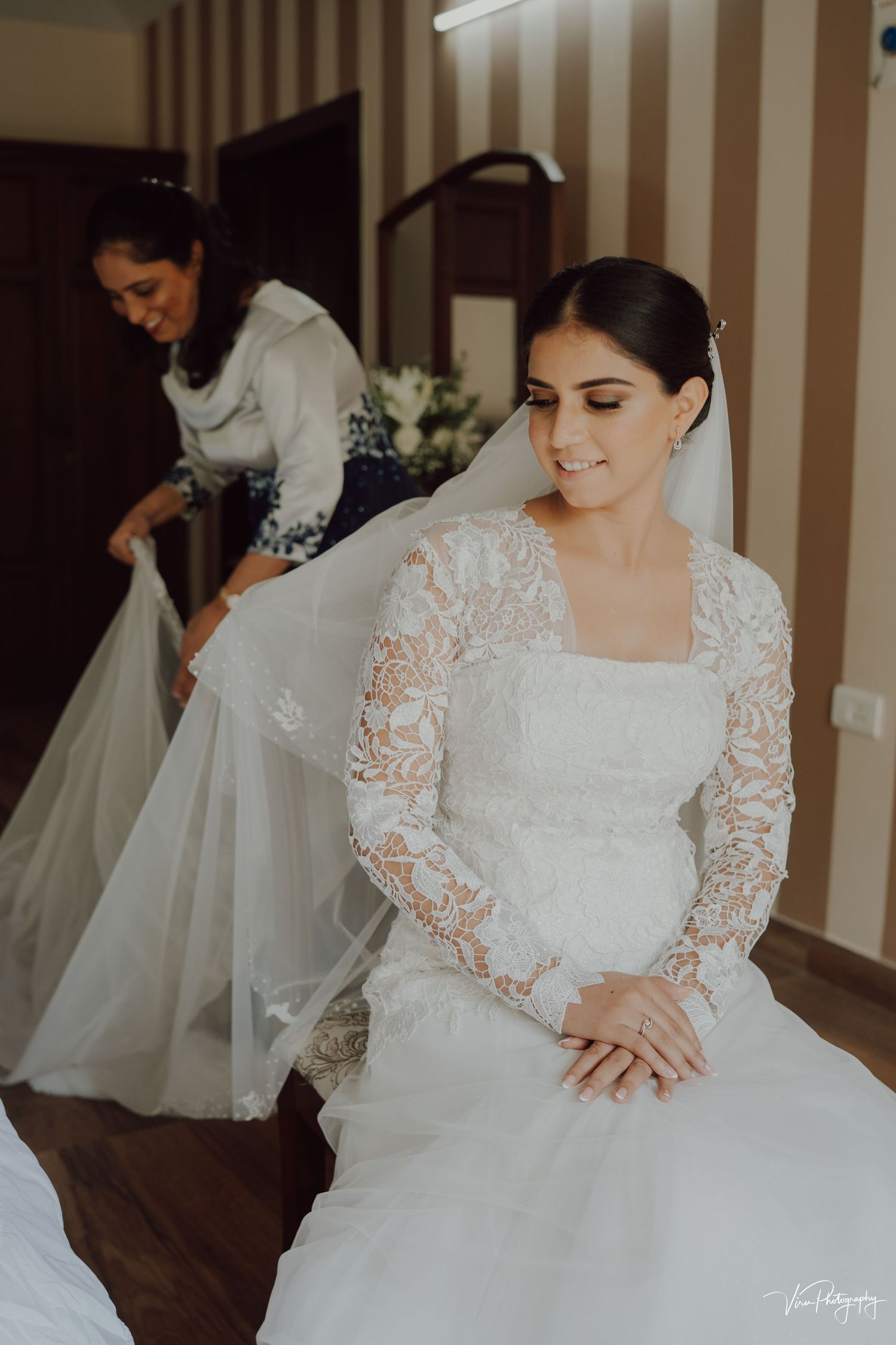 A bride in a long-sleeved lace wedding dress sits calmly while her gown is being adjusted in a striped-wall room.
