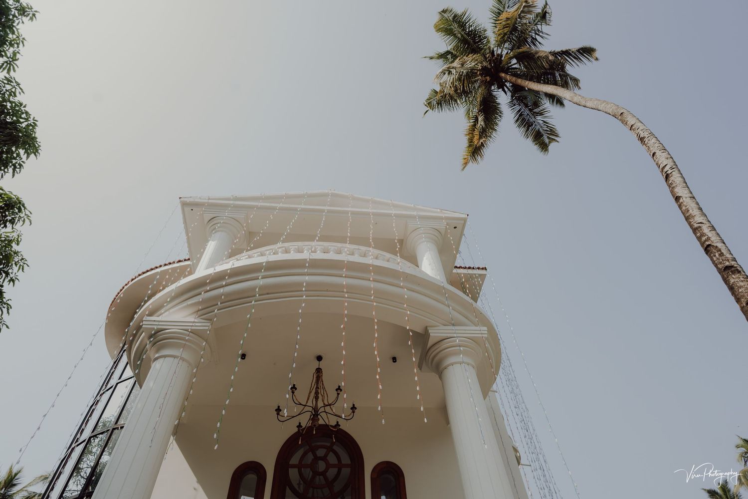 White church entrance with a chandelier and tall curved palm tree against a cloudy sky in tropical setting.