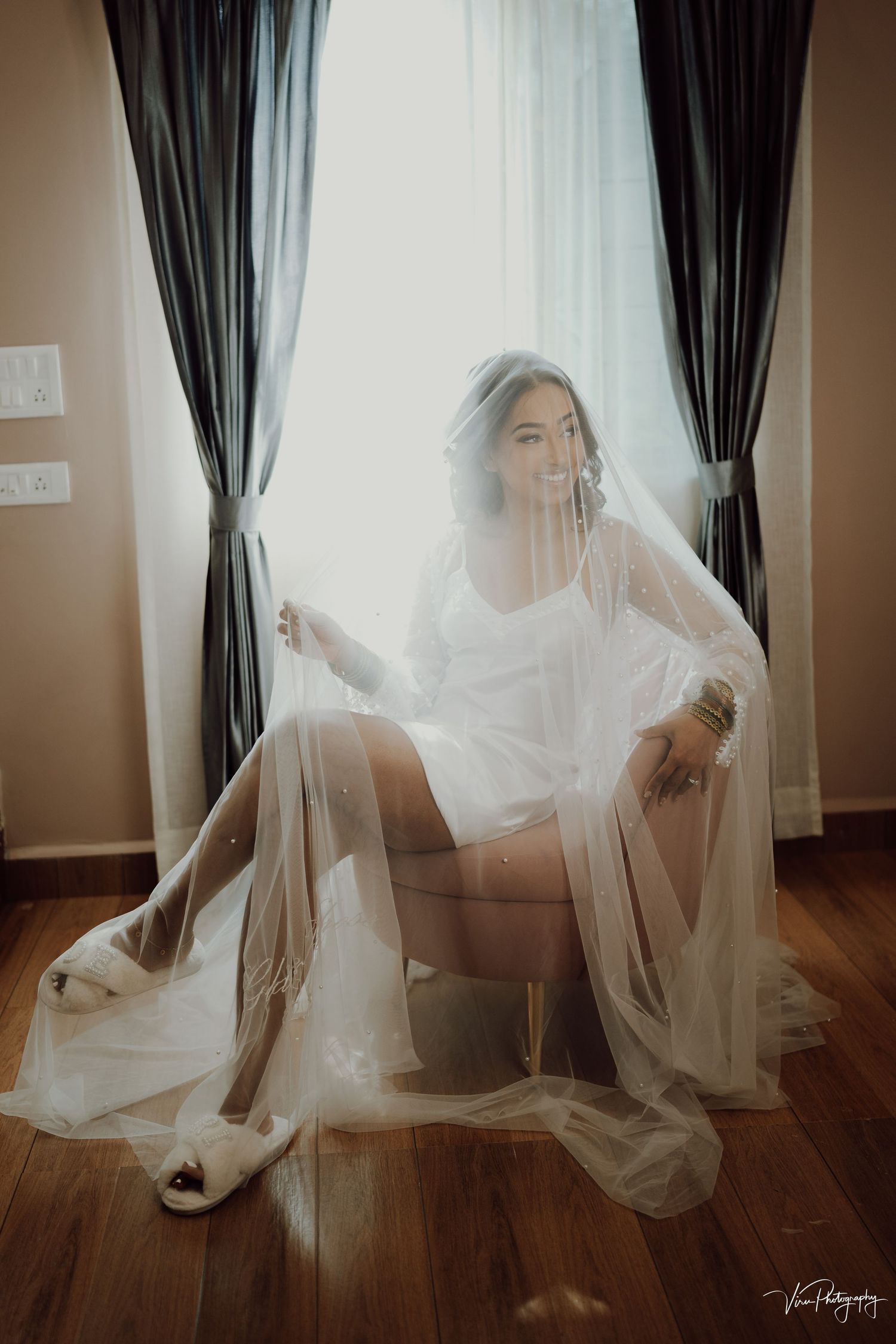 A person in a white bridal robe and flowing veil sits elegantly on a chair near curtained windows with soft natural light.