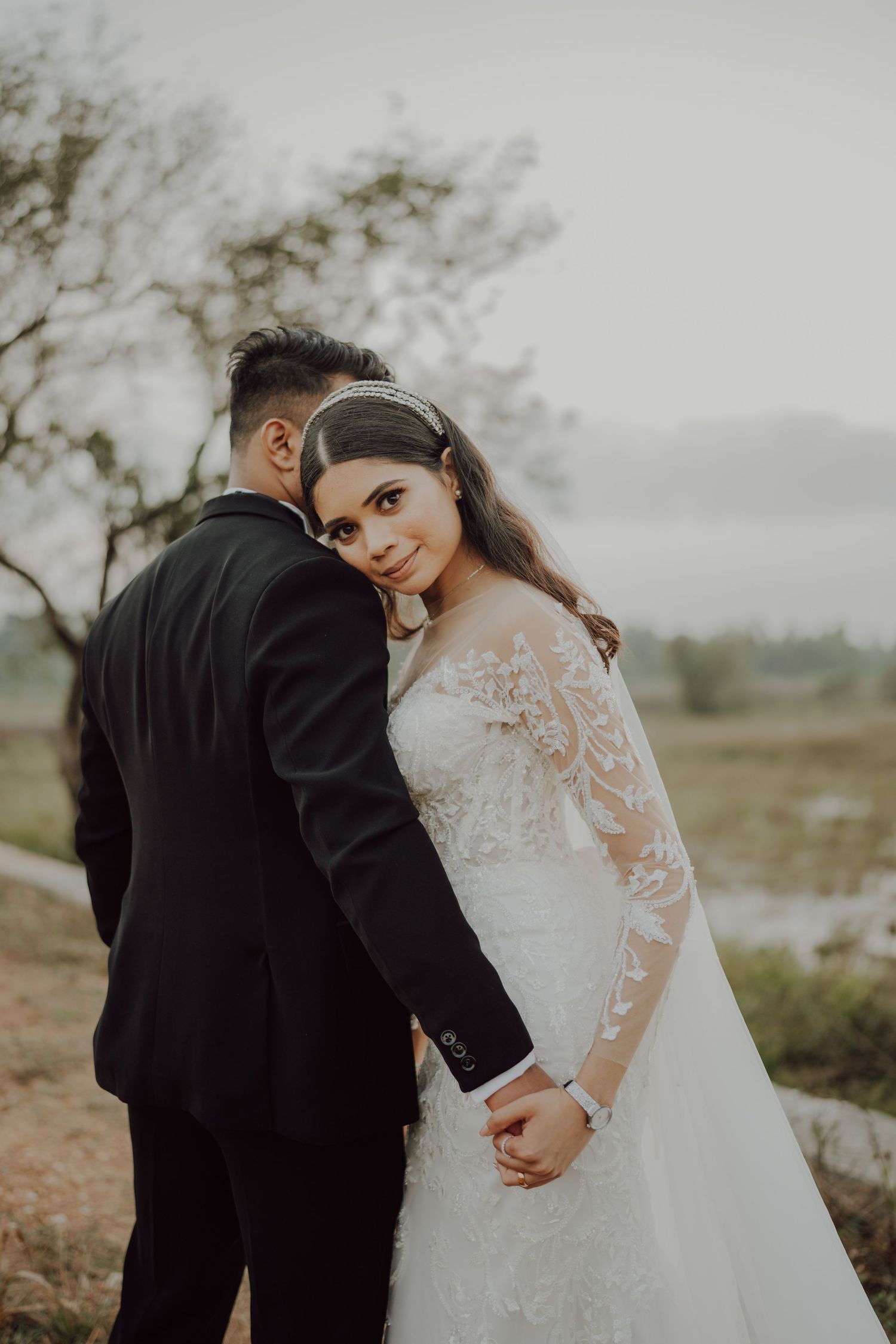 A bride in a lace sleeve wedding gown stands with her groom in black suit against a misty outdoor backdrop.
