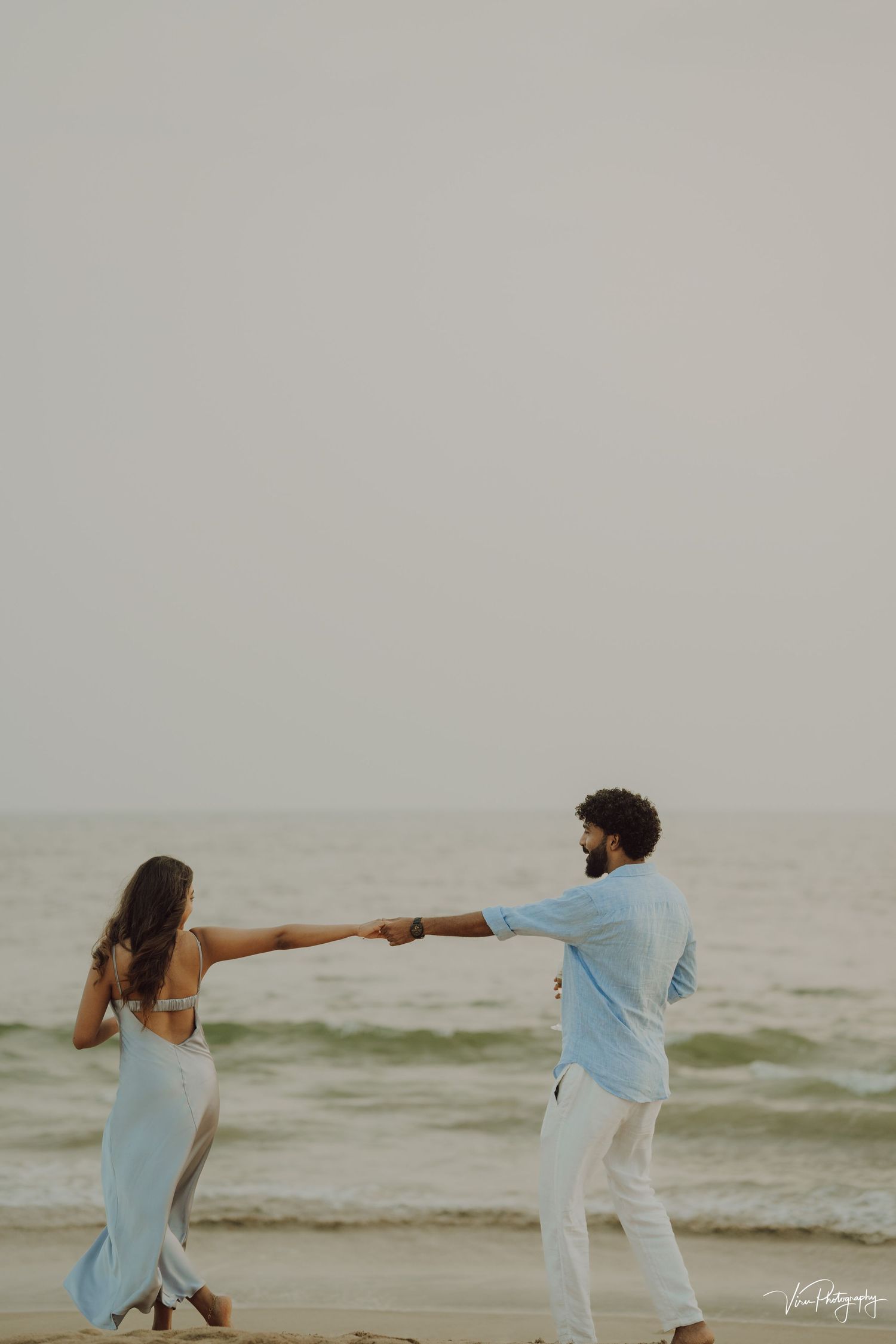A couple holds hands and dances together on a misty beach at sunset, wearing flowing light blue and white outfits.