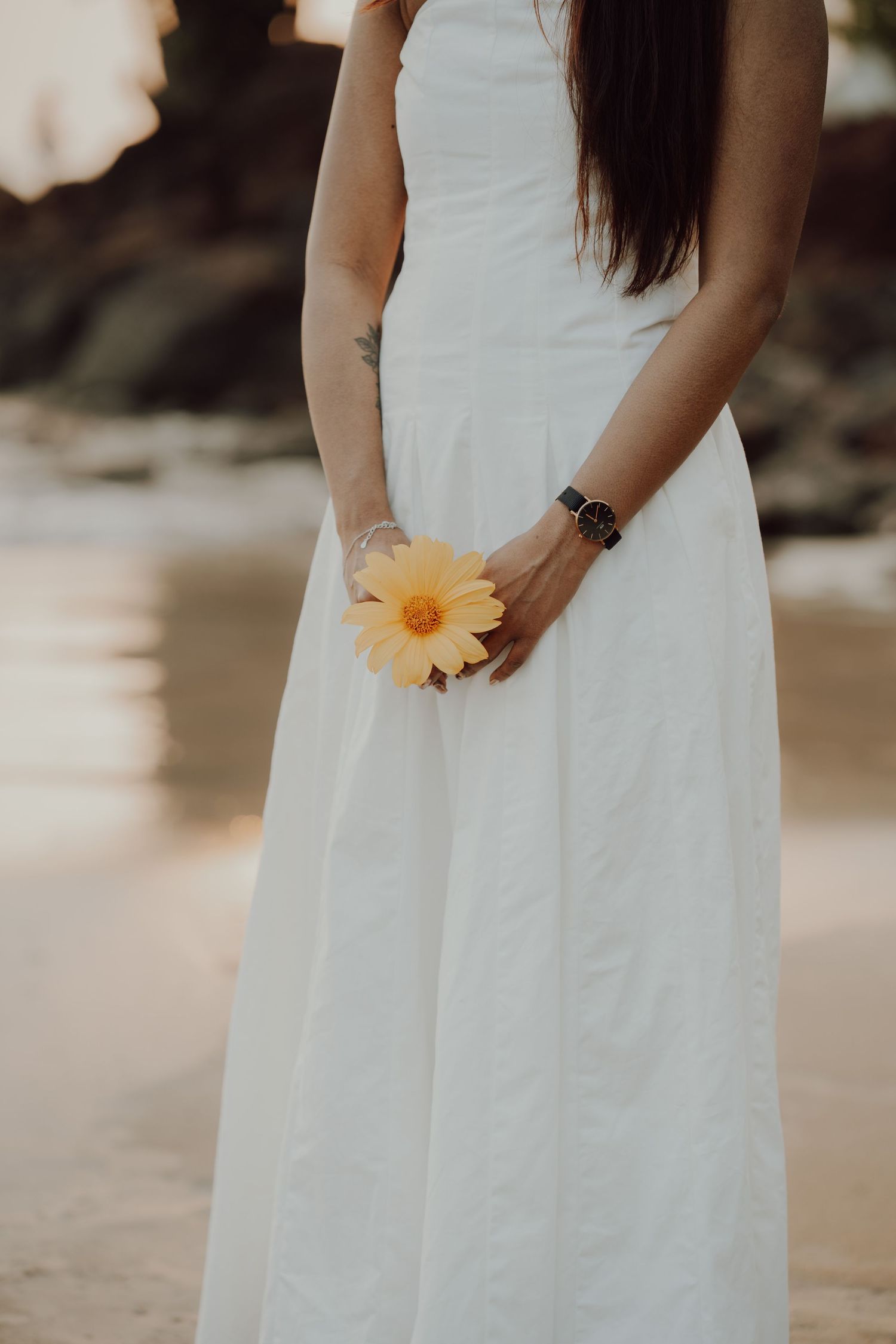 A person in a flowing white dress holds a single yellow daisy flower on a beach at sunset.