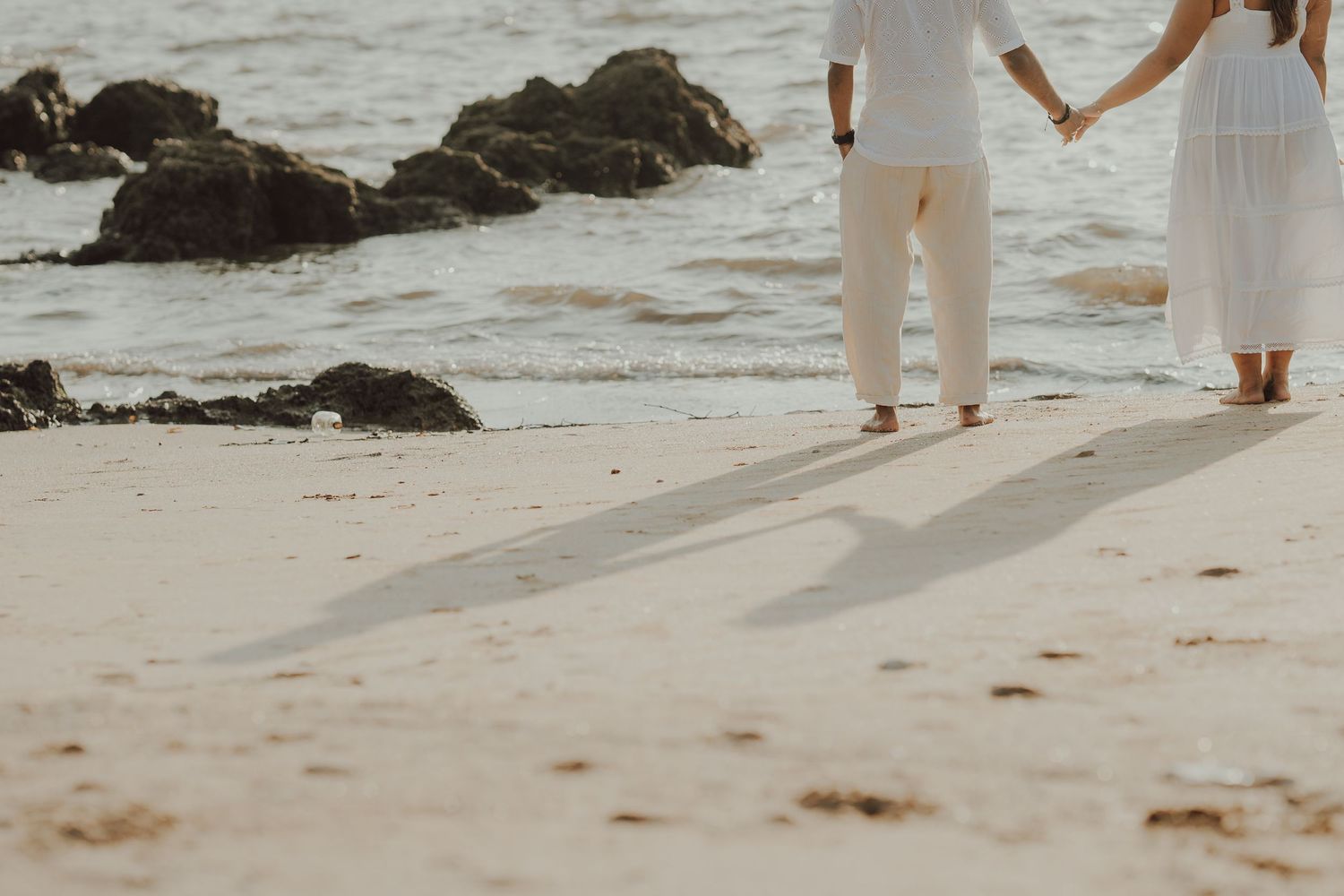 People in white outfits walking along a rocky beach shoreline at sunset casting long shadows on the sand.