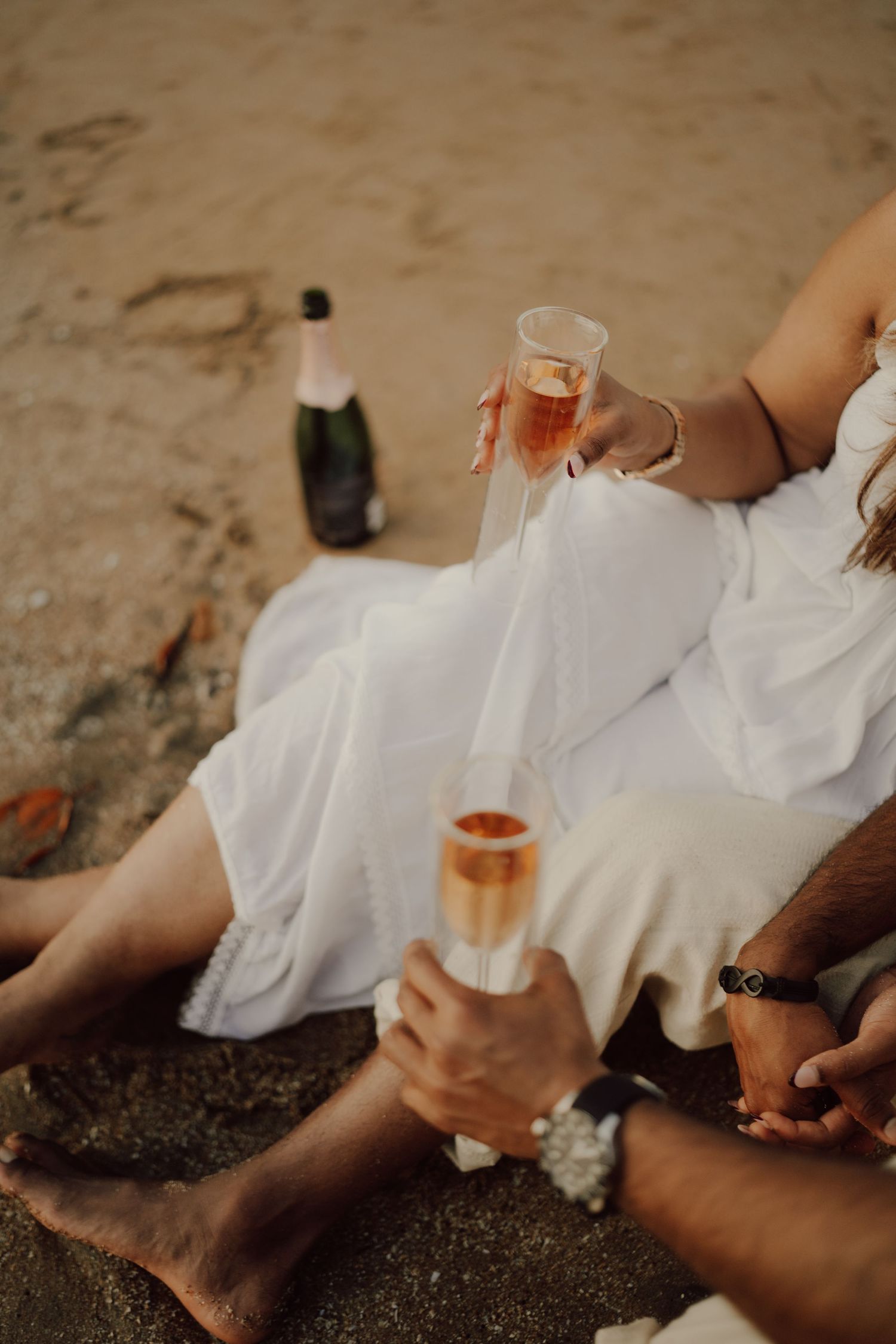 Two people in white clothing sit on beach sand holding glasses of rose wine at sunset.