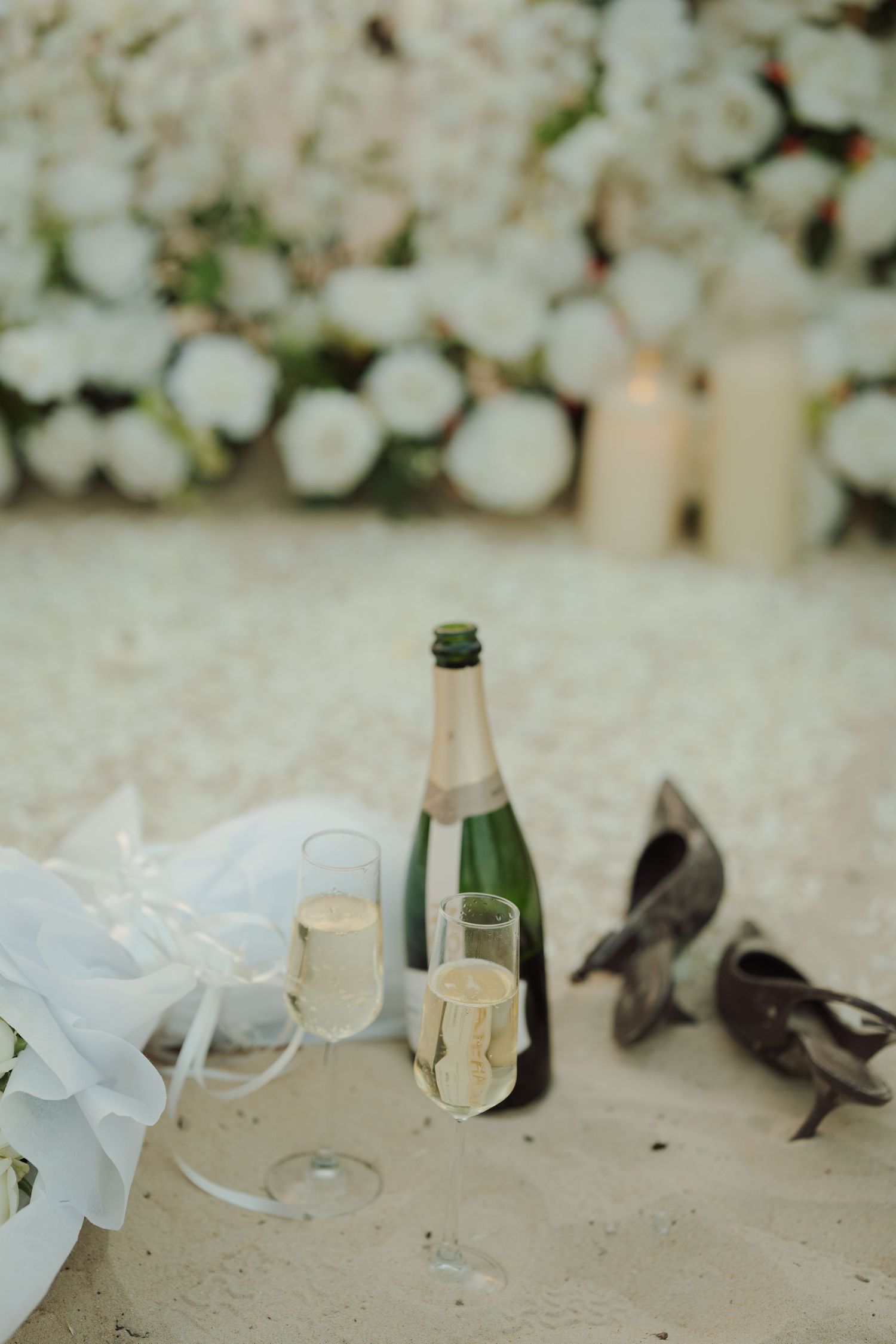 Champagne bottle with two glasses on sandy beach, white floral backdrop, heels and bridal bouquet nearby.