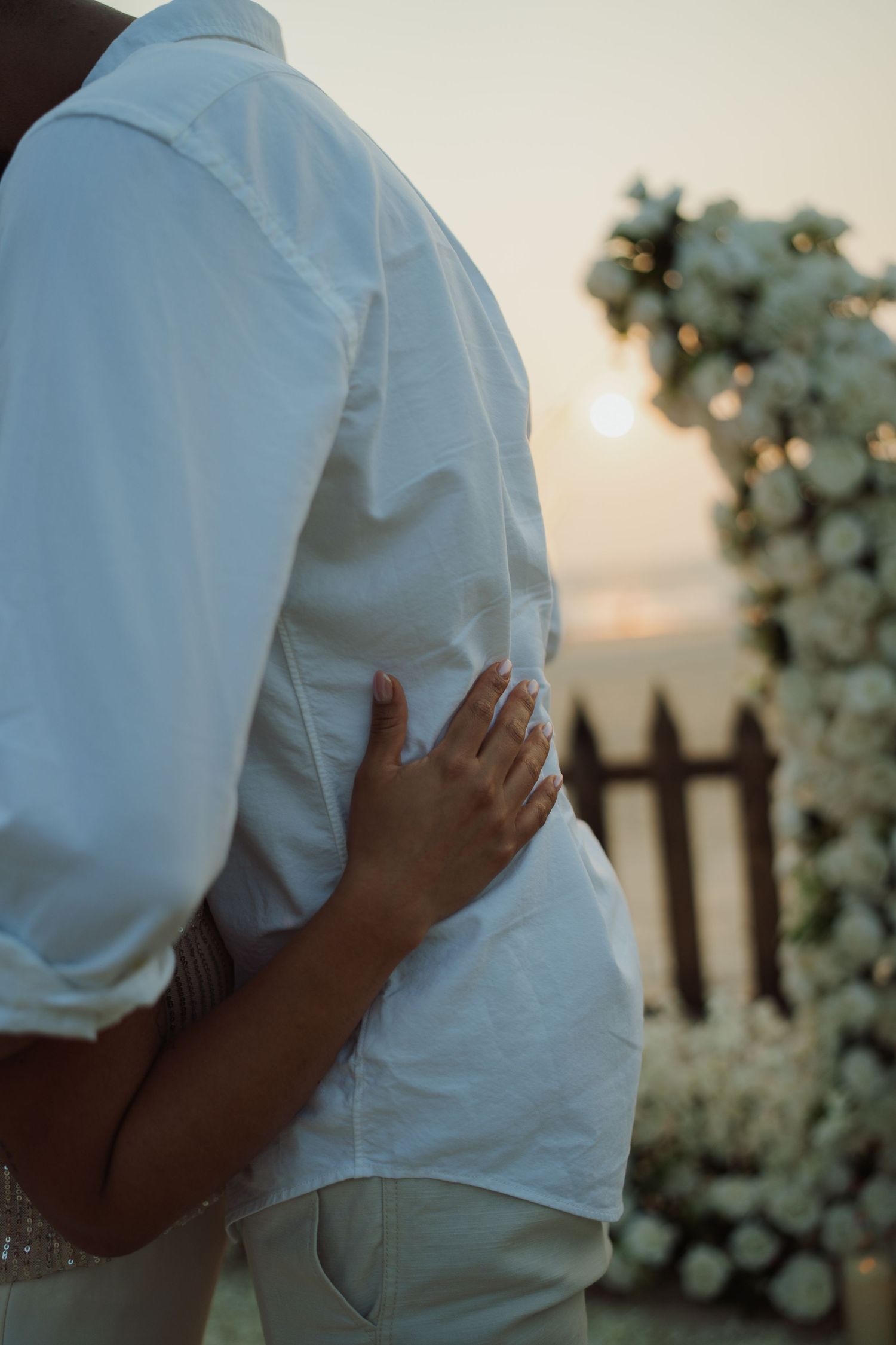 Couple embracing at sunset near a floral arch, woman wearing a ring, both dressed in white.