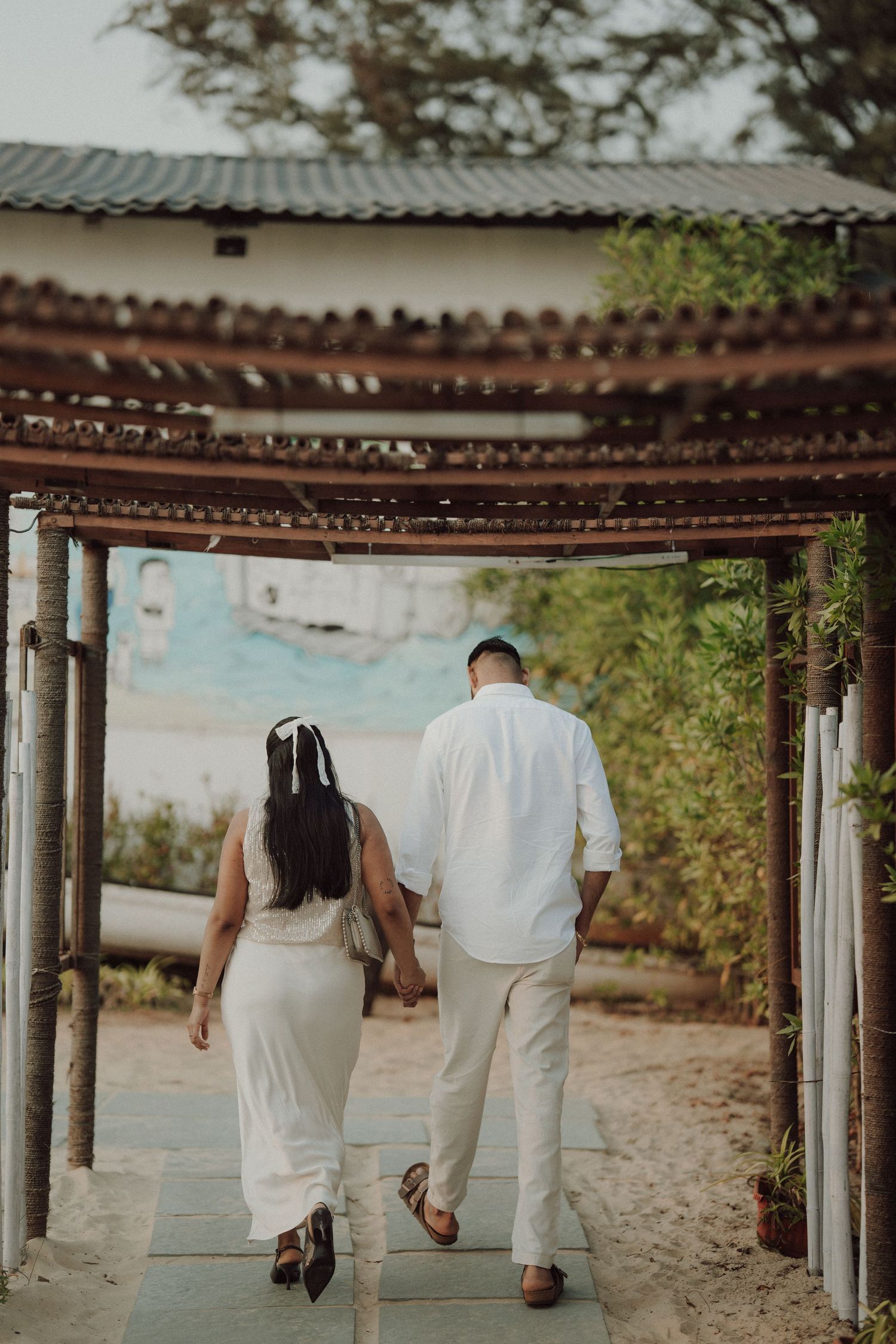 Couple in white outfits holding hands walking under a rustic wooden pergola toward a tropical beach setting.