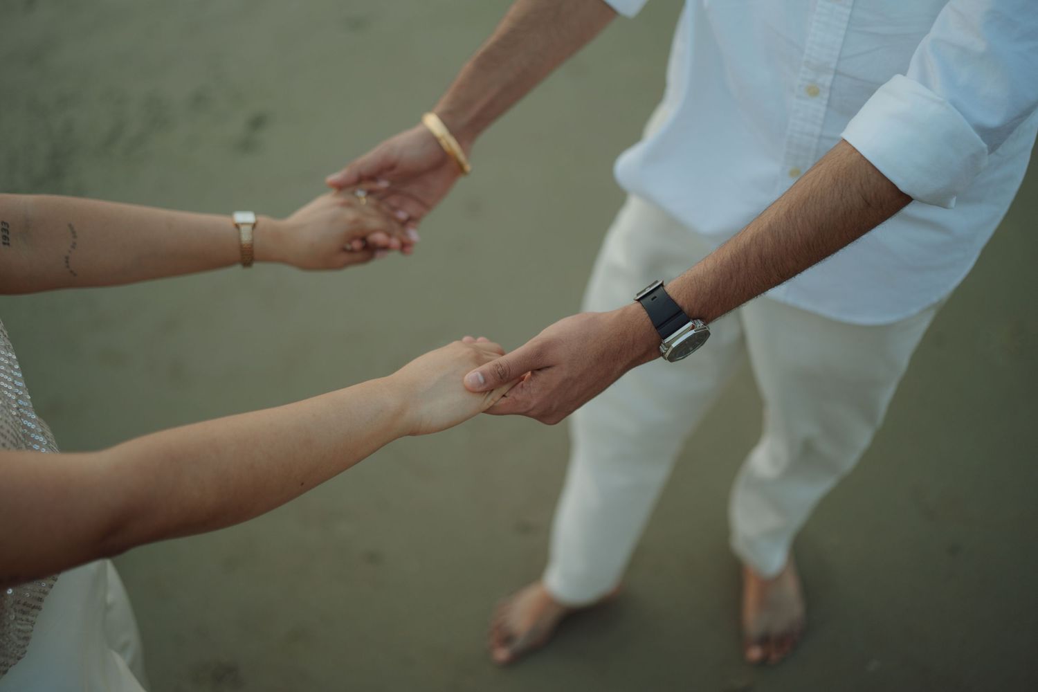 Couple holding hands on a beach, wearing white outfits with a bracelet and smartwatch visible.