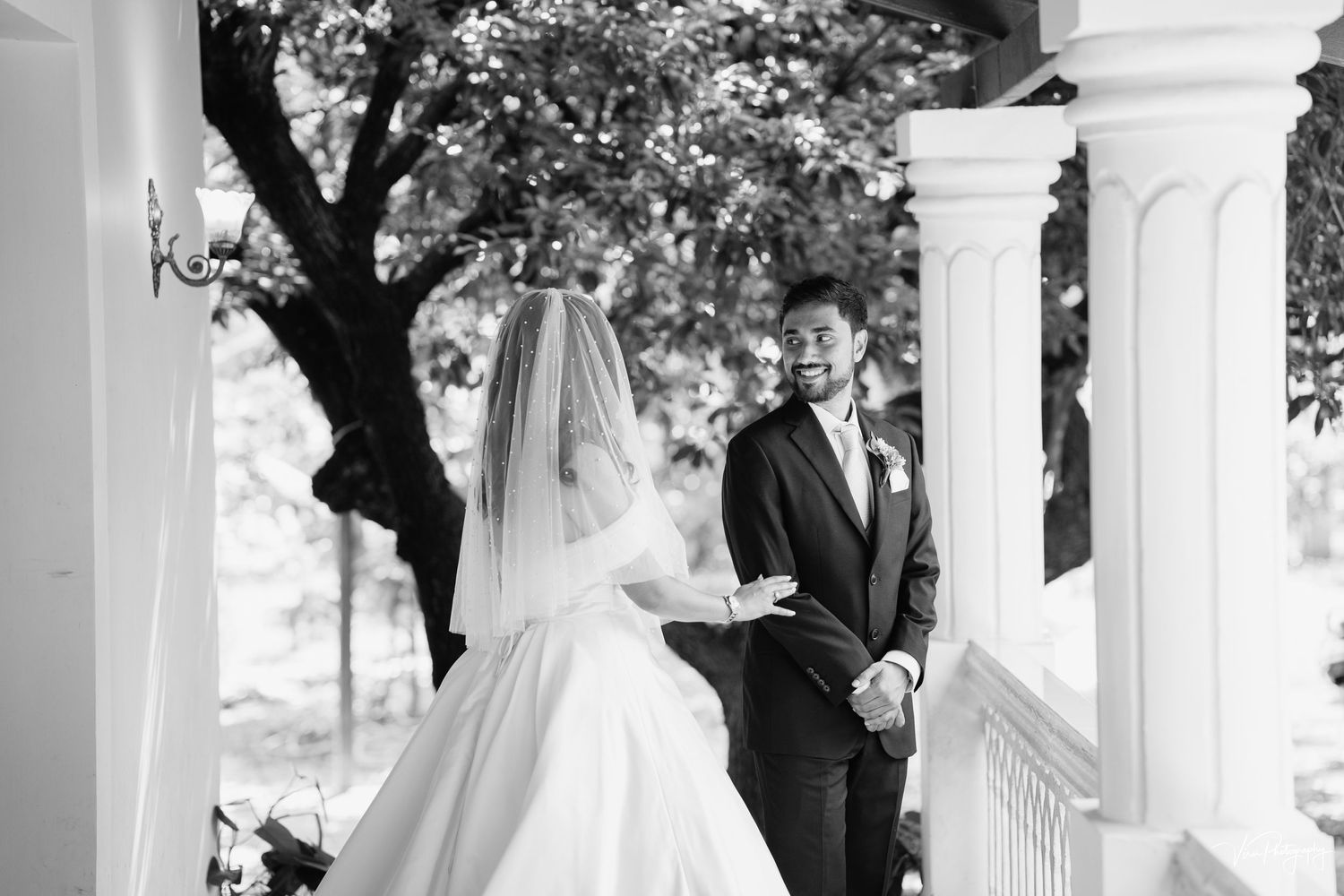 A couple in traditional black and white wedding attire share a moment on a white columned porch.