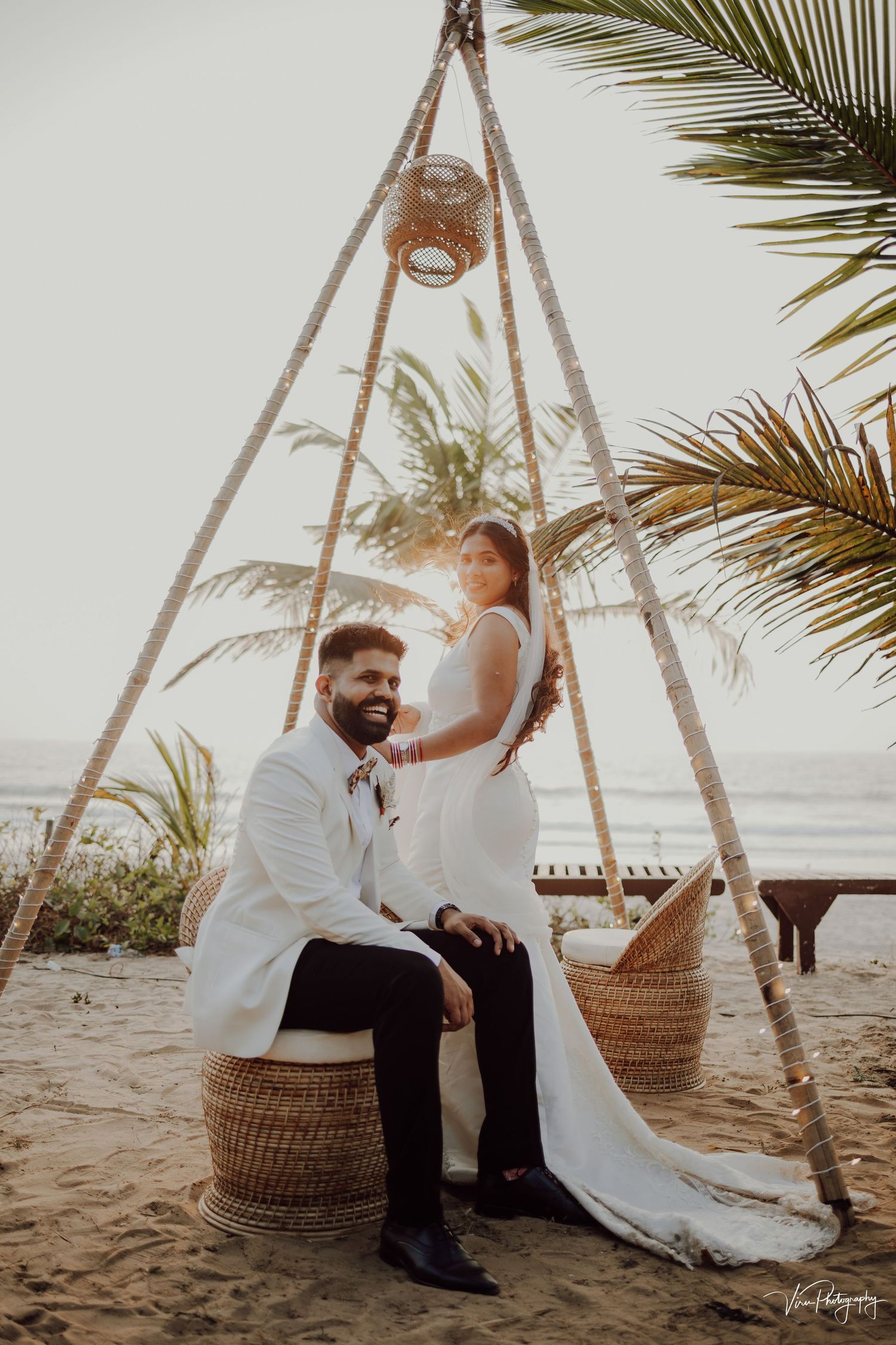 A beachy boho wedding setup with a triangular arch, woven seating, and palm trees beside the ocean at sunset.
