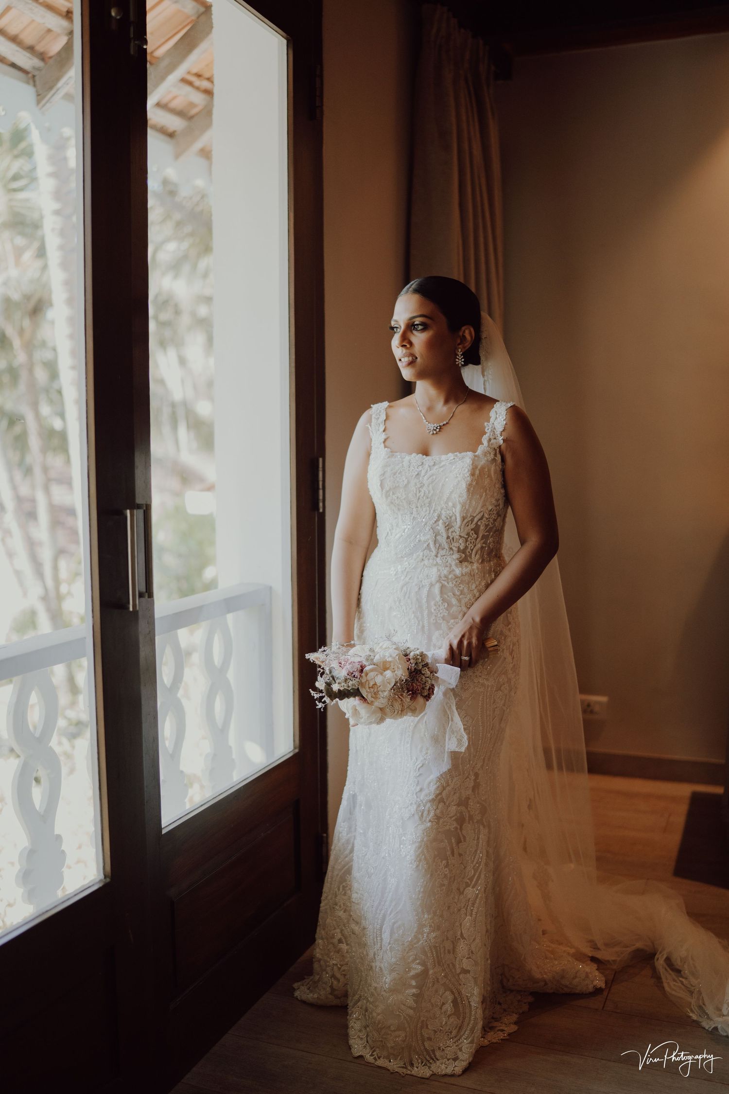 A bride in a lace wedding gown stands elegantly by a window in warm light, holding a delicate bouquet.