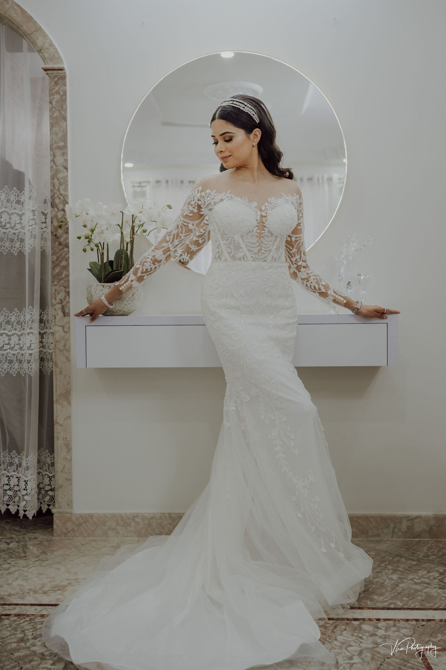 Elegant long-sleeve lace wedding gown with illusion neckline and fitted mermaid silhouette shown against circular mirror.