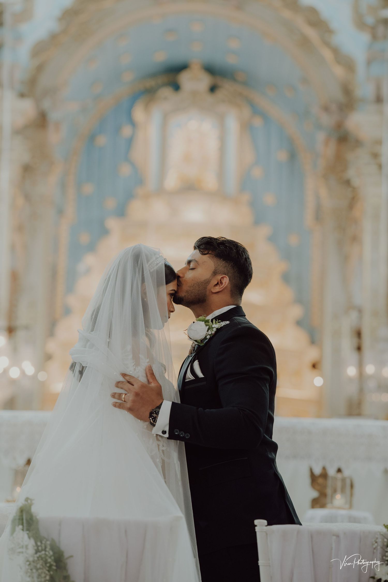 Newlyweds share romantic kiss against ornate baroque church altar with beautiful soft lighting and flowing white veil.