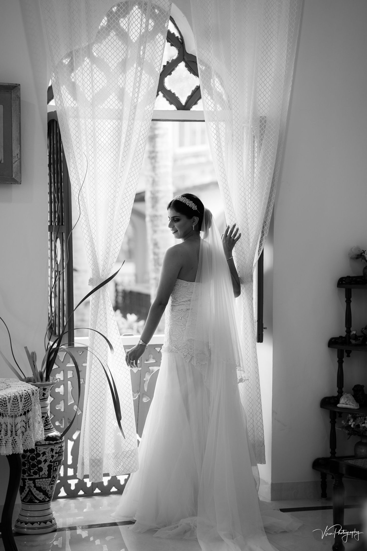 Elegant black and white photo of a bride in flowing wedding dress and veil standing by a window with sheer curtains.