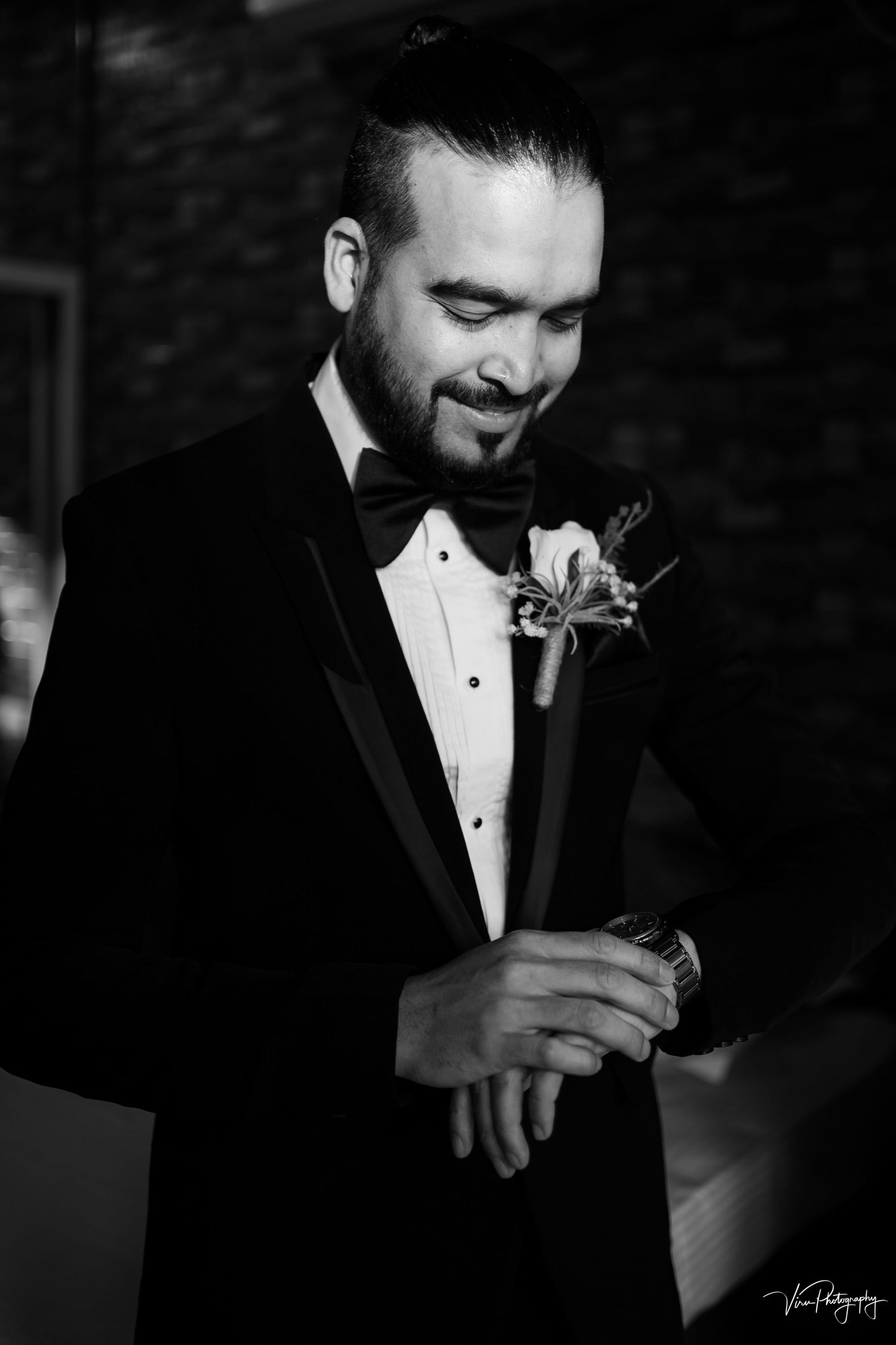 A groom in a black tuxedo and white boutonniere smiles while looking down in this black and white portrait.