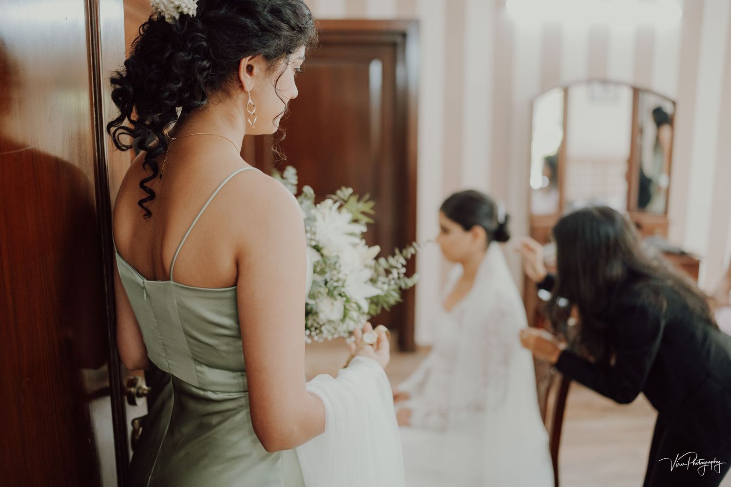 Bridesmaid in sage green dress holds bouquet while watching bride get ready in dressing room.