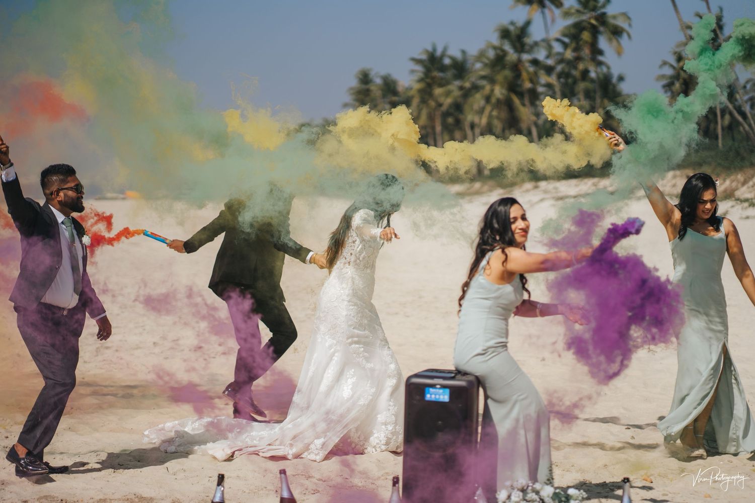 Wedding party celebrates with colorful powder explosions on a beach during a joyful dance sequence.