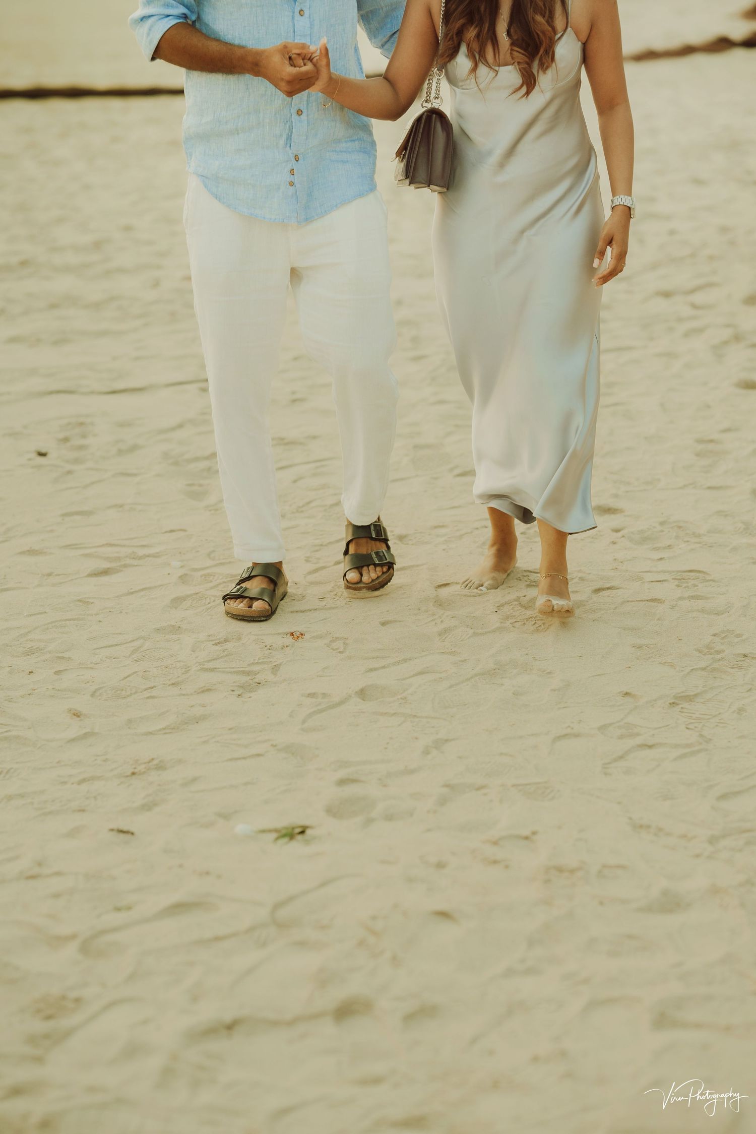 A couple walking barefoot and in sandals on a sandy beach wearing casual white outfits.