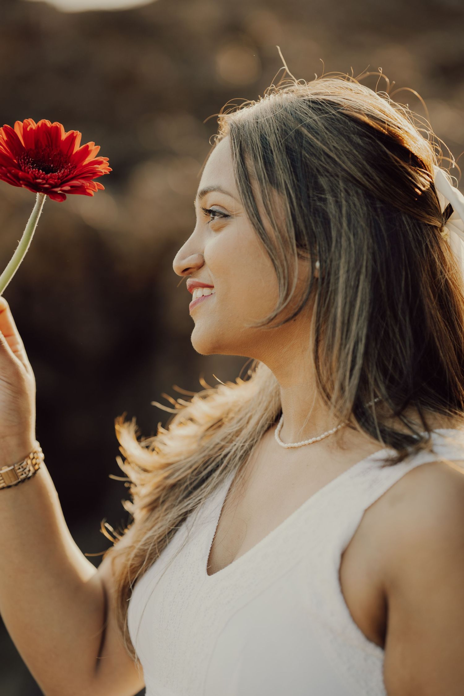 A woman in a white dress holds up a red gerbera daisy flower against a warm sunset backdrop.