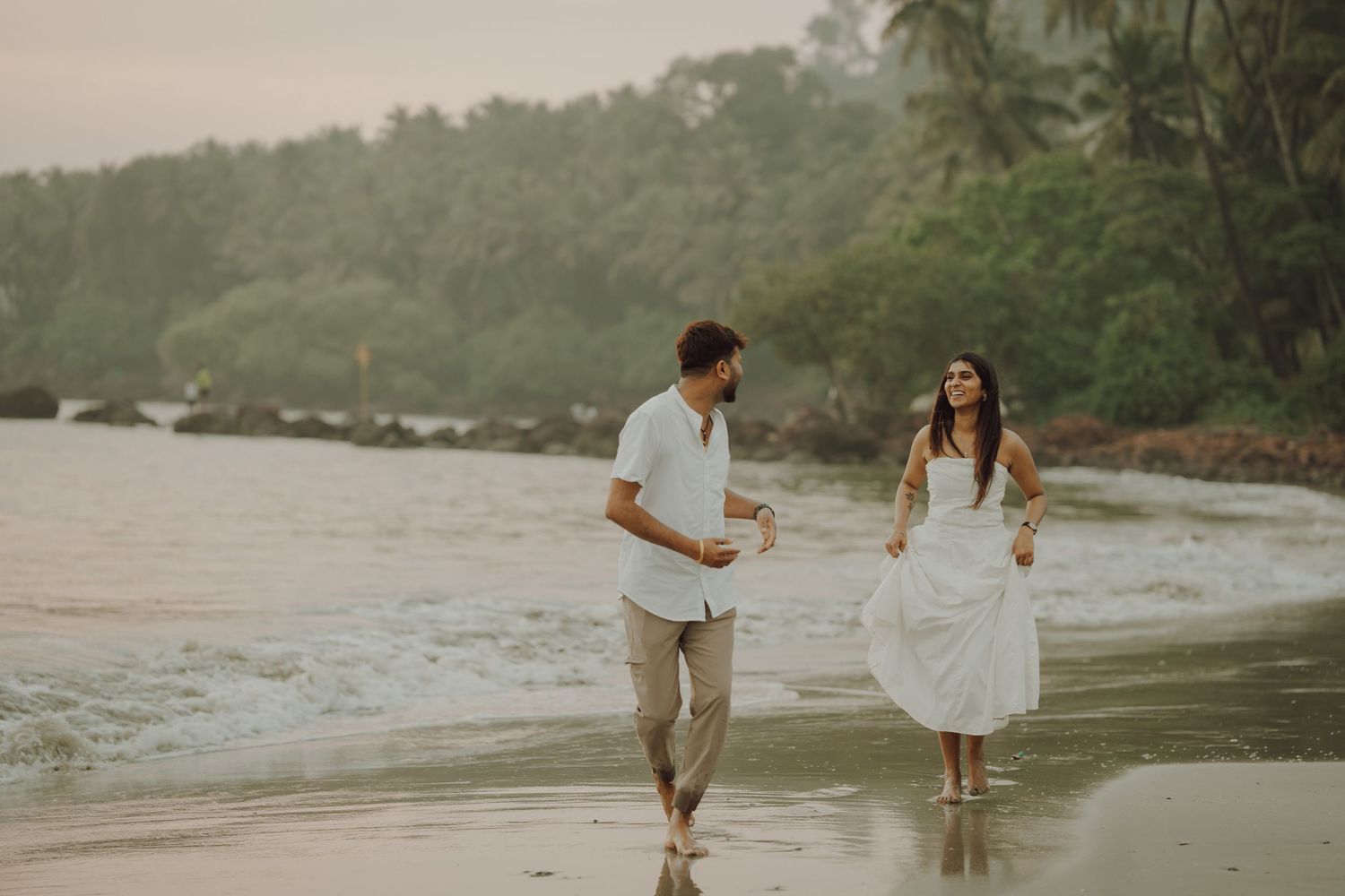 A couple walks hand in hand along a misty tropical beach at sunset creating a romantic silhouette against the shoreline.