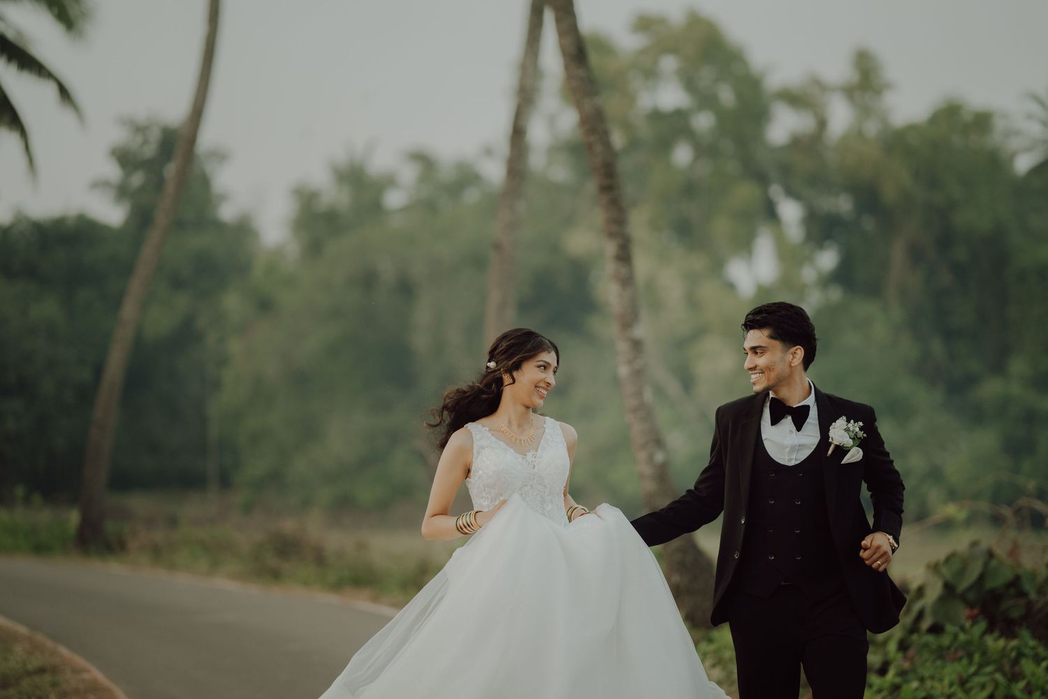 Wedding couple walking hand in hand on palm-lined road with bride in flowing white gown.
