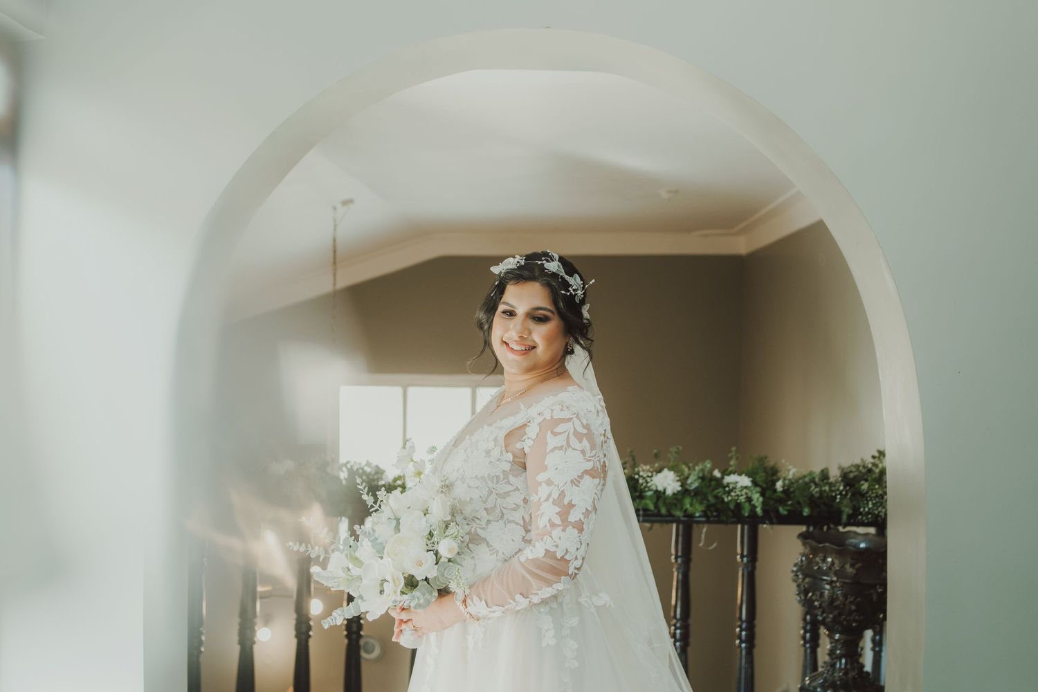 Side view of bride in white lace gown holding bouquet under arched doorway.