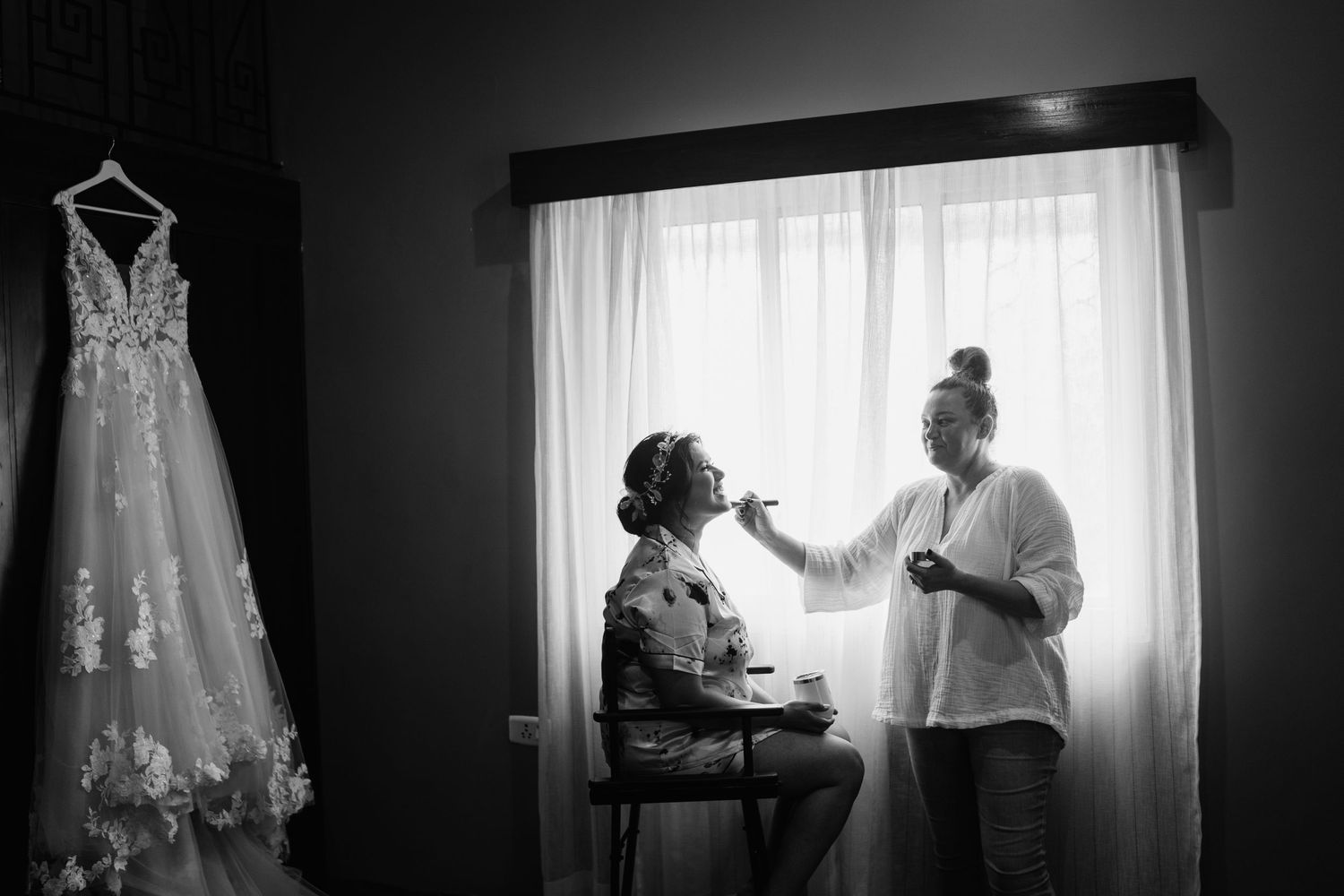 Black and white image of bridal preparation with wedding dress visible in background.