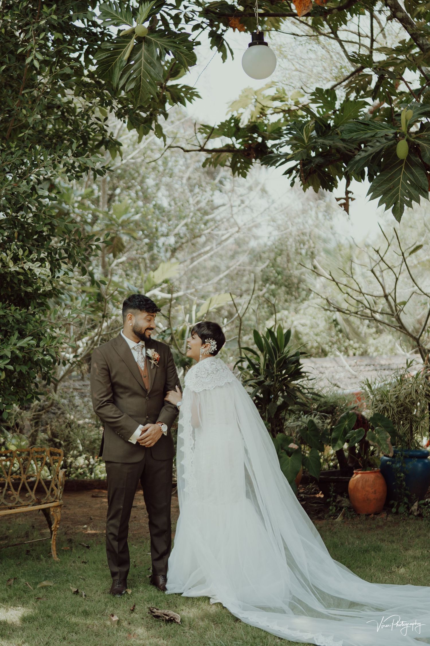A couple shares an intimate moment in a garden wedding setting with lush greenery and a flowing white cape dress.