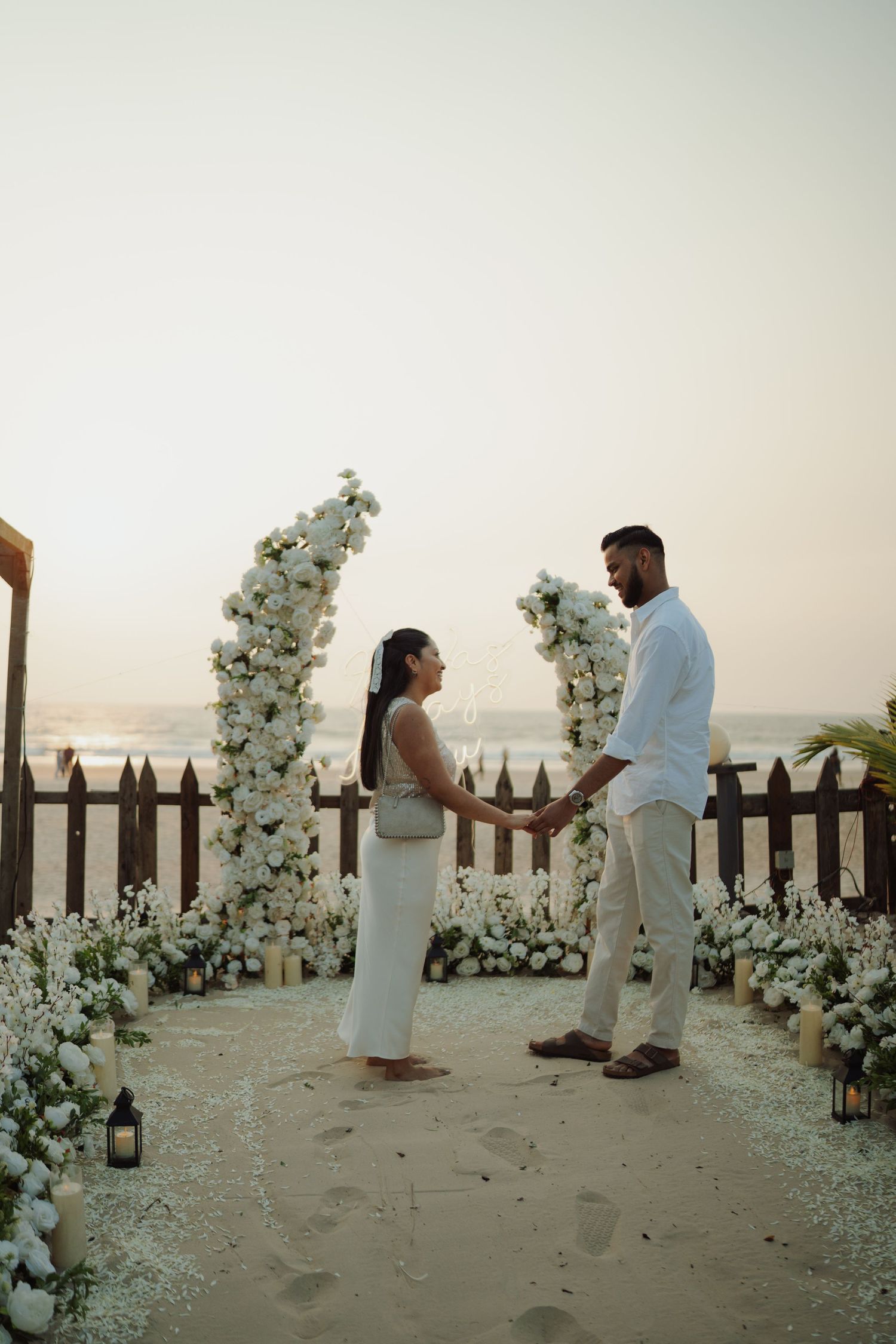 Couple holding hands at a romantic beach wedding ceremony with white floral arch and lanterns at sunset.